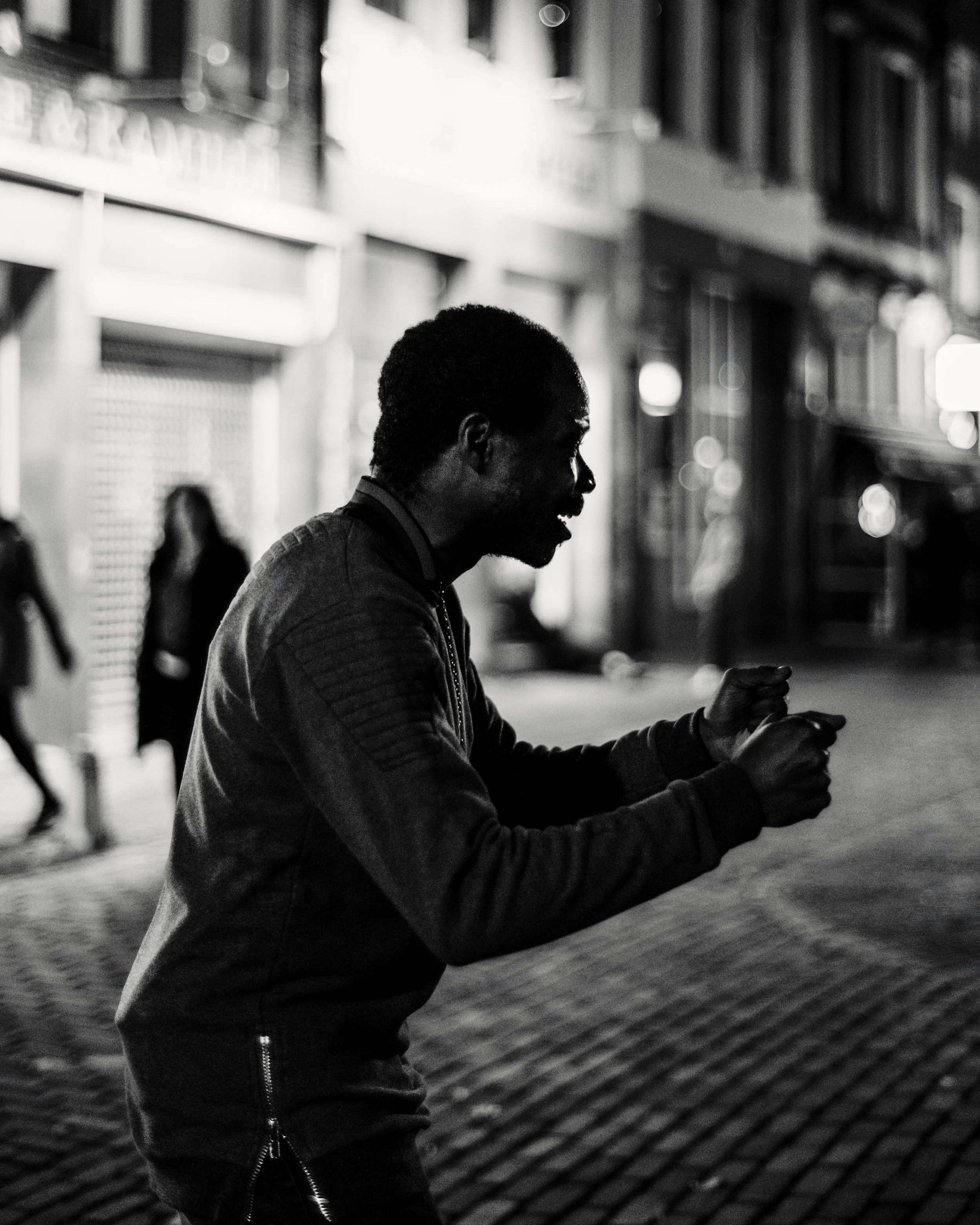 Black and white image of a person with curly hair, wearing a jacket, sitting outdoors at night, looking at their phone with a smile.