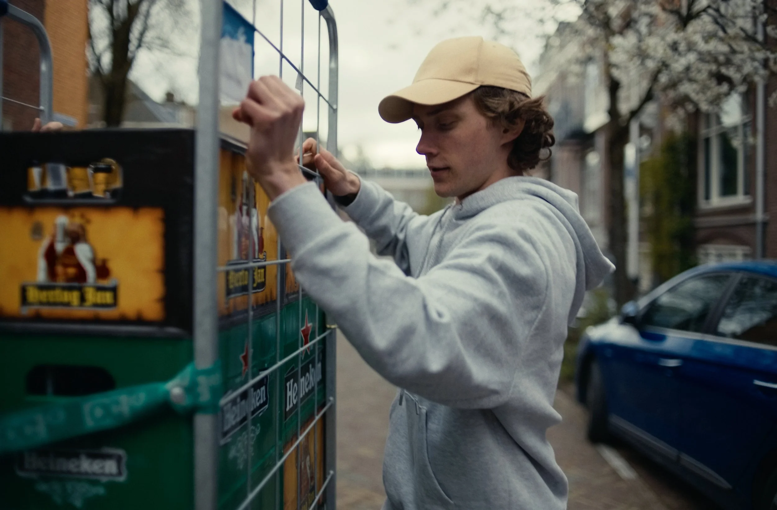 A young man with curly hair, wearing a beige cap and a light gray hoodie, is buying snacks from a vending machine outdoors in a residential area with trees and parked cars.