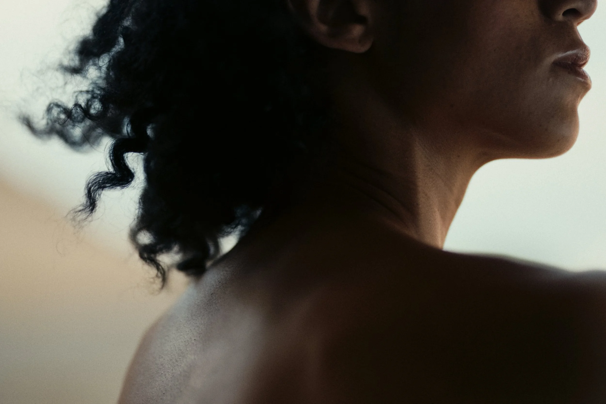 Close-up side profile of a woman with dark wavy hair and smooth skin, with a neutral background.