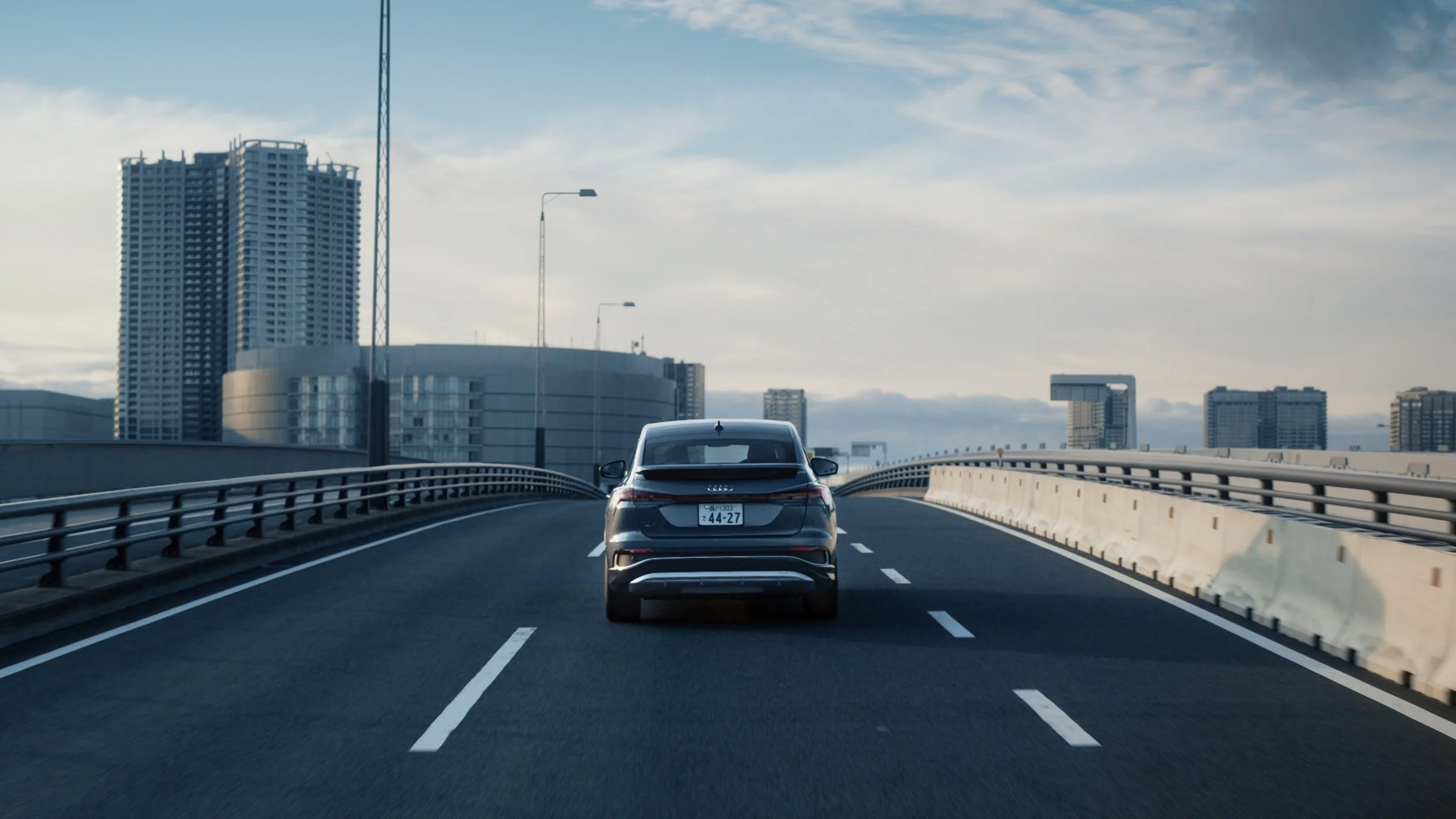 A black sedan driving on an elevated highway with city skyscrapers in the background and a partly cloudy sky.