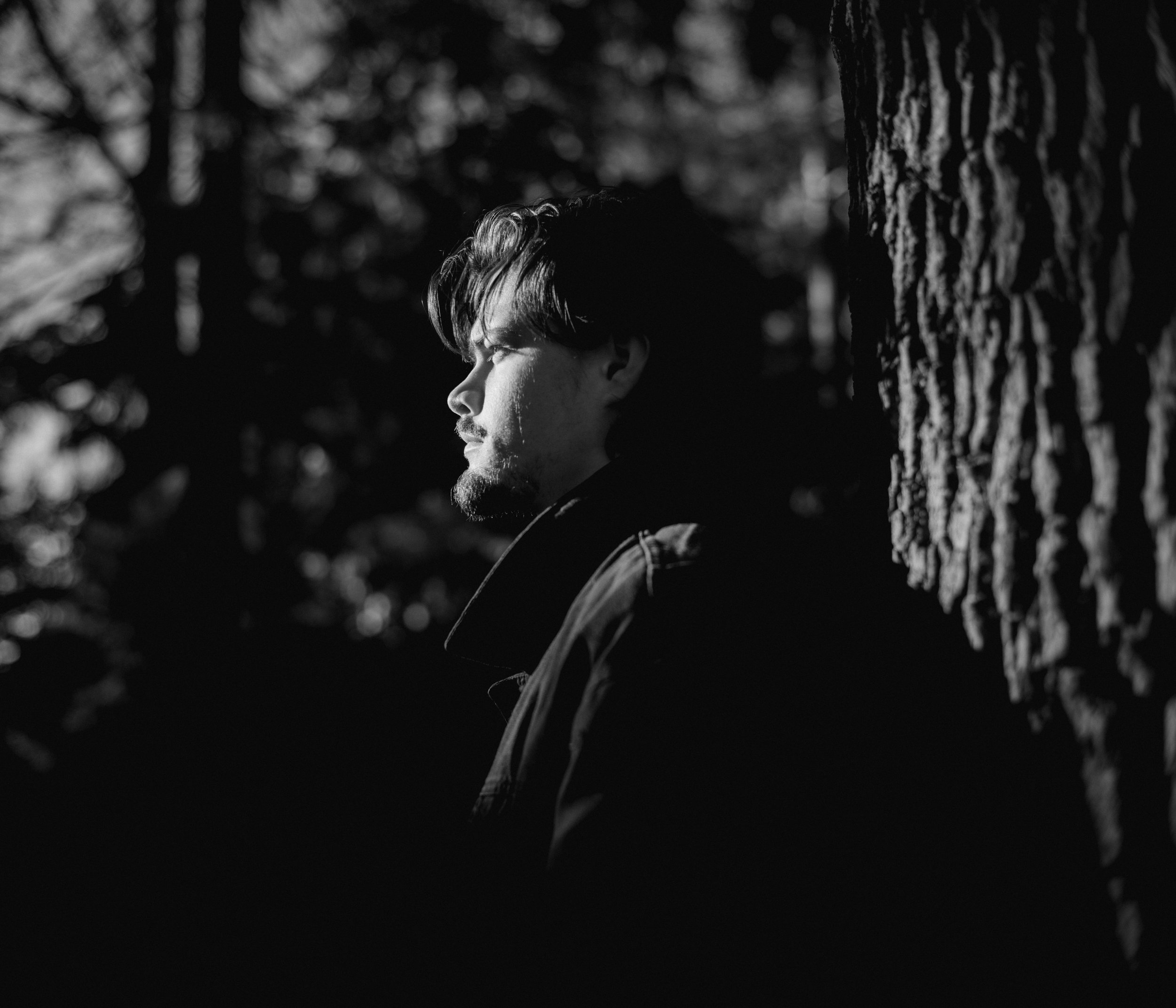 A black and white photograph of a person with light skin and dark hair, leaning against a tree outdoors. The person's face is in profile, and they appear to be looking into the distance with a thoughtful expression. The background consists of blurred