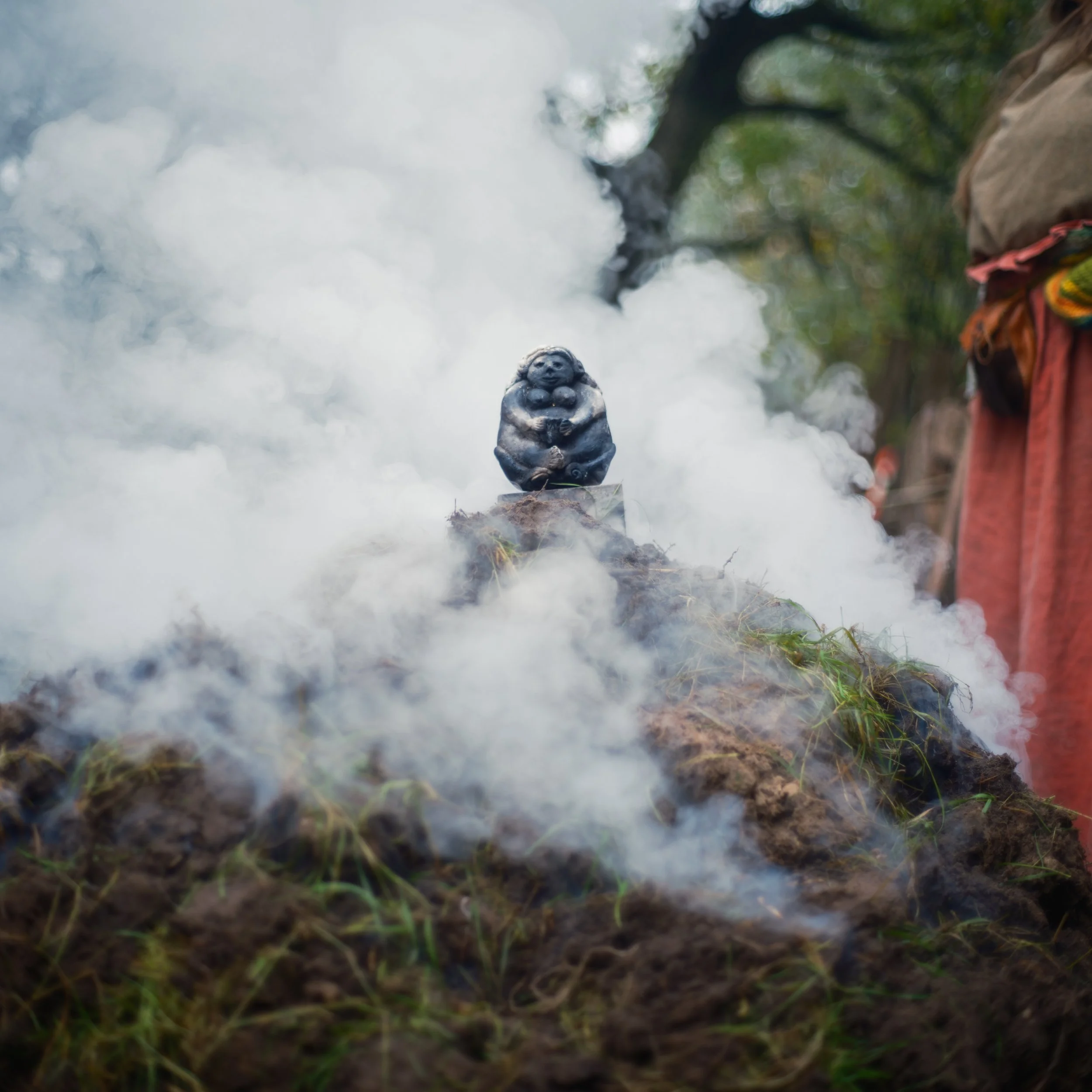 A small, dark-colored figurine or sculpture of a figure, possibly a mythical or spiritual character, is situated on the ground amidst fog, with trees and greenery in the background.