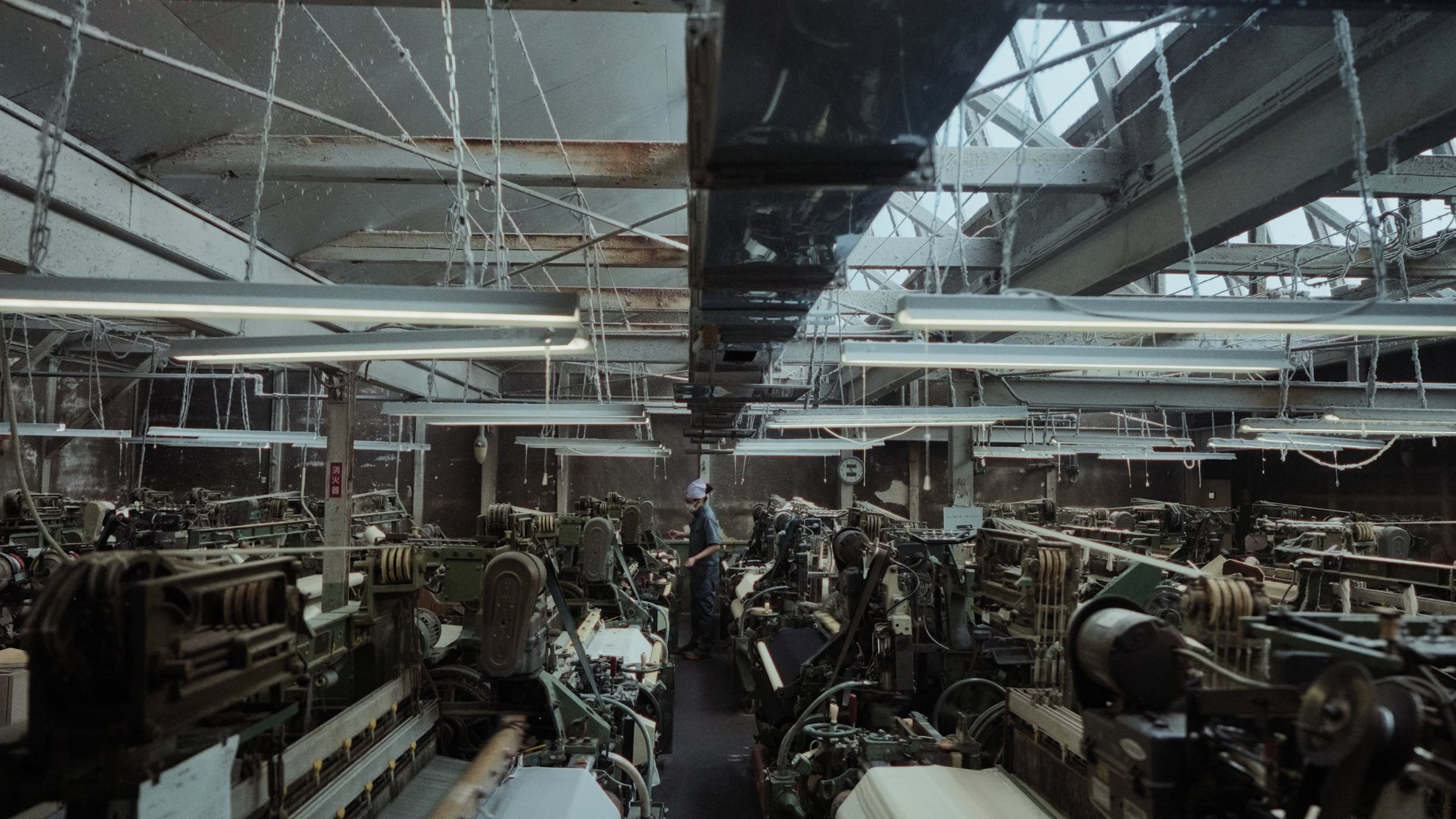 A factory or workshop filled with machinery and equipment, with a worker inspecting or operating the machines, surrounded by hanging wires and industrial lighting.