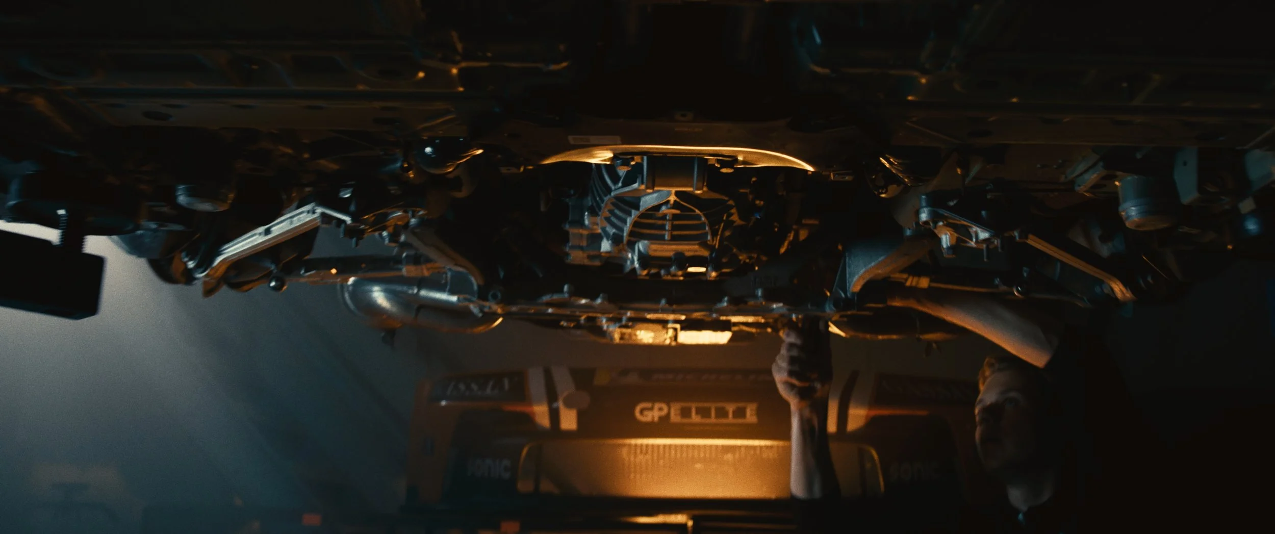 A person standing in front of a large mechanical structure, possibly on a film set, with a vehicle labeled "G-PELLI" in the background. The scene has a dark, cinematic atmosphere with warm lighting.