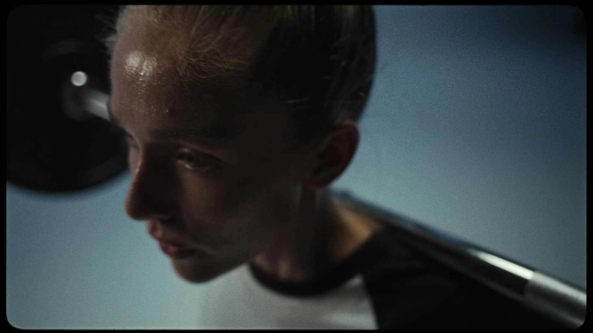 A close-up of a man with blonde hair and sweat on his forehead, looking downward in a dimly lit room.