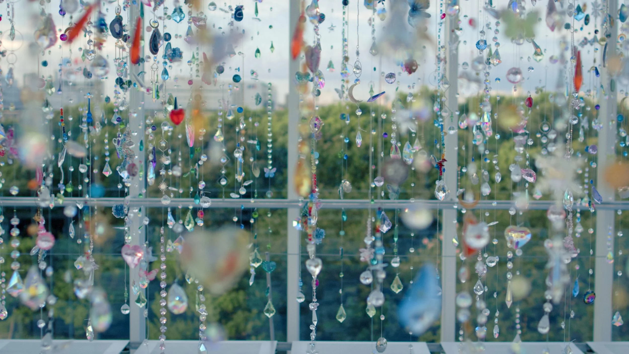 A collection of hanging crystal ornaments and beads arranged vertically in front of windows, with a view of trees outside in the background.