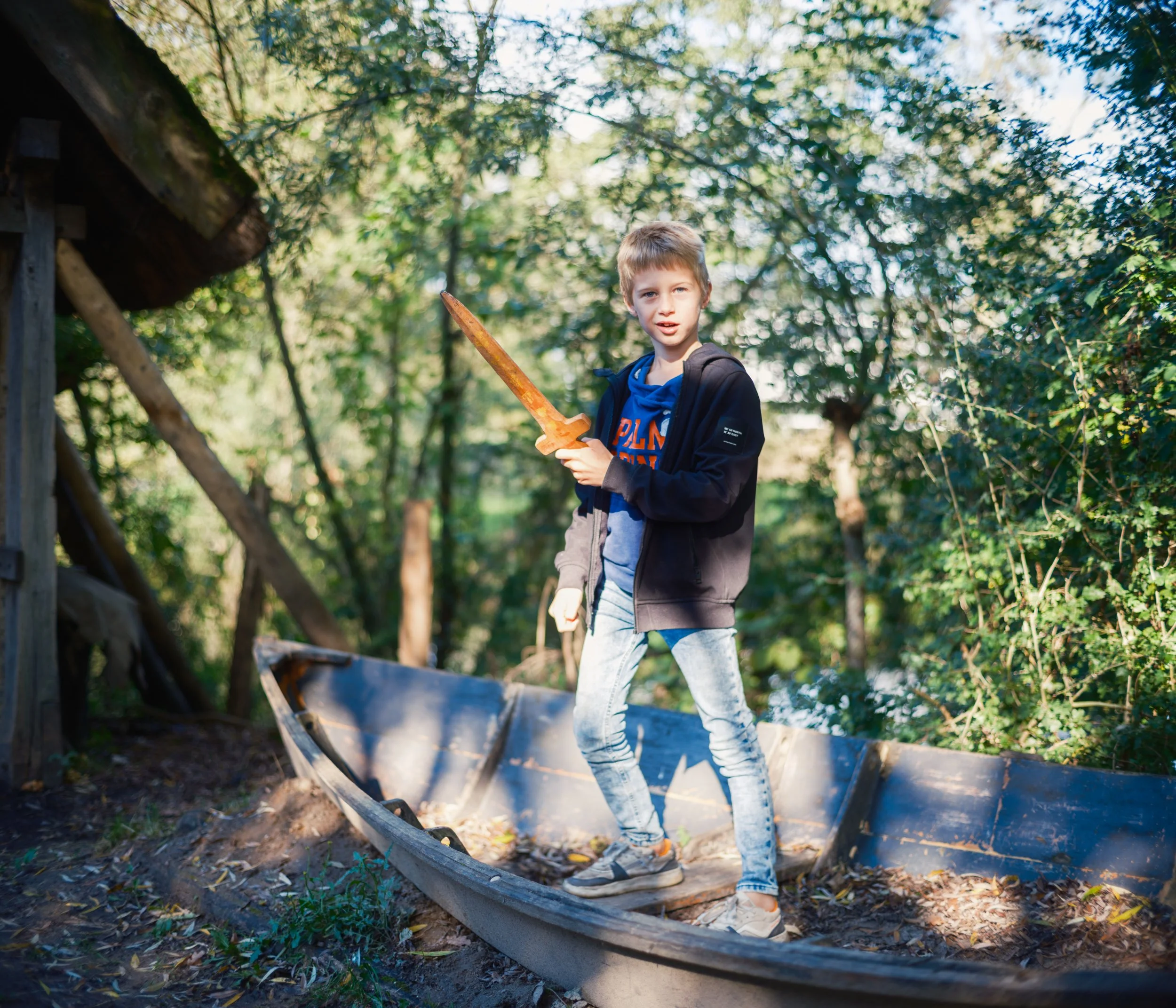 A young boy with light brown hair, wearing a black jacket and blue hoodie, holding a small orange toy sword while standing in an outdoor wooden boat in a wooded area with green trees and sunlight.