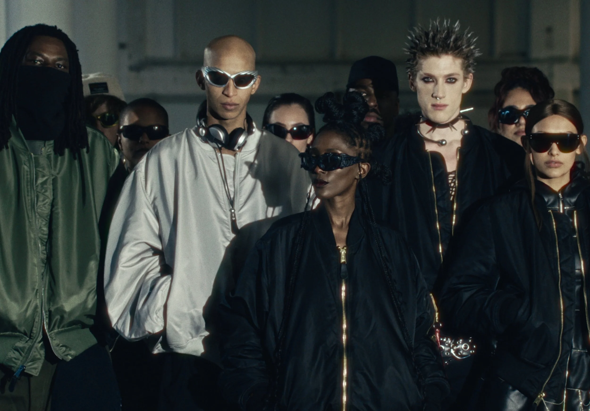 A group of diverse young people wearing fashionable clothing and accessories, standing together in a dimly lit setting.