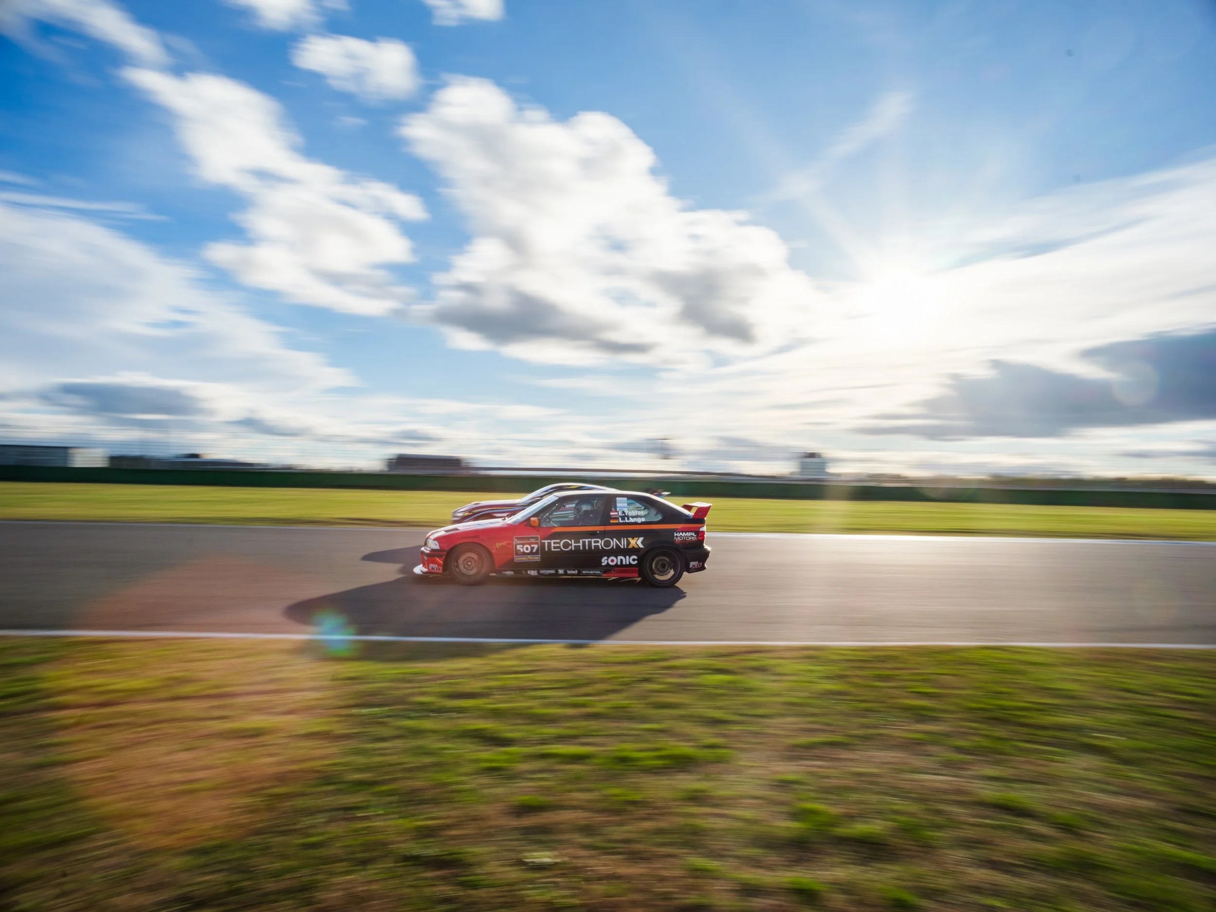Race car driving on a race track during daytime with a bright blue sky and scattered clouds.