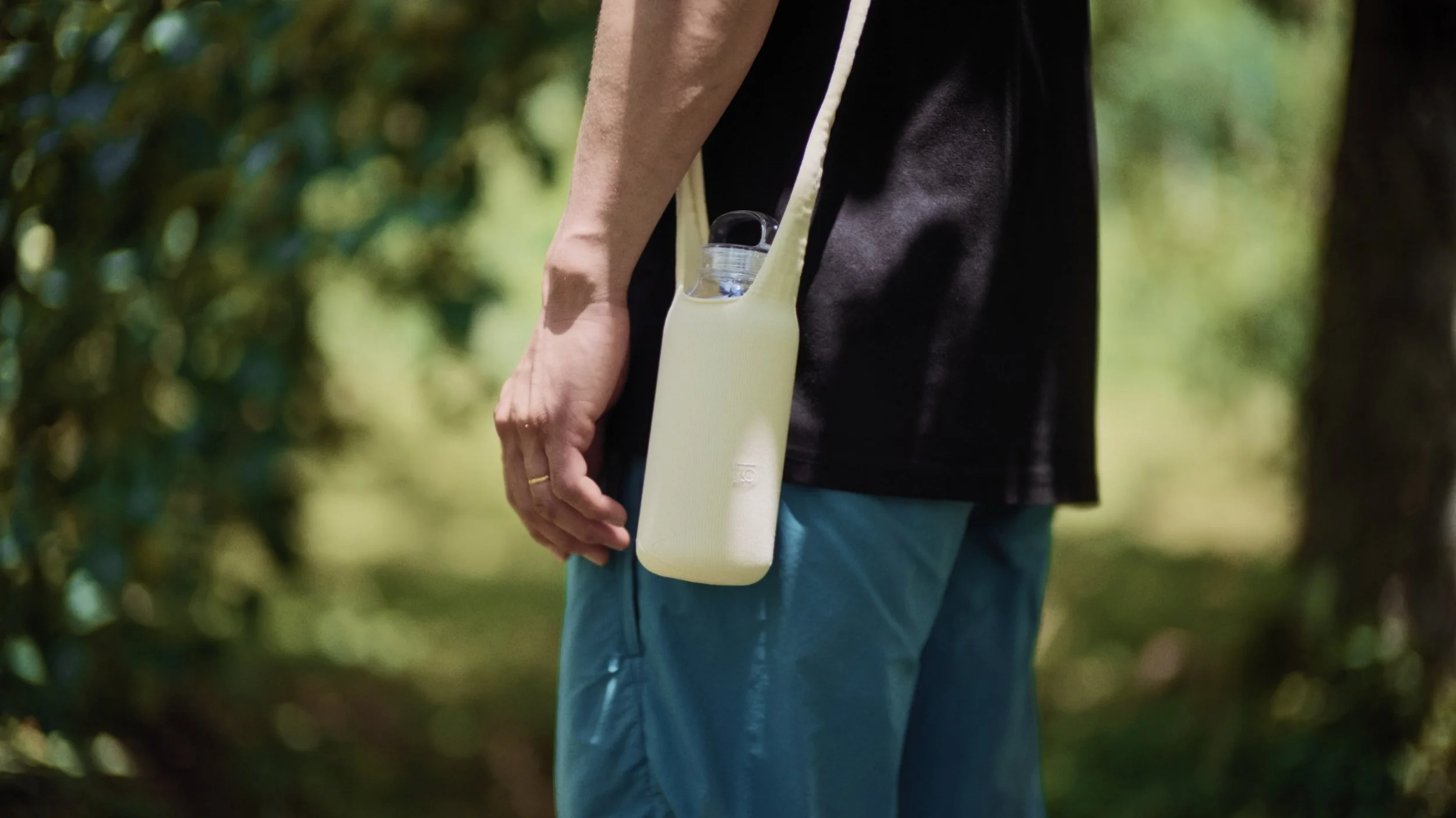 Person holding a water bottle with a strap around their neck, outdoors in a wooded area.