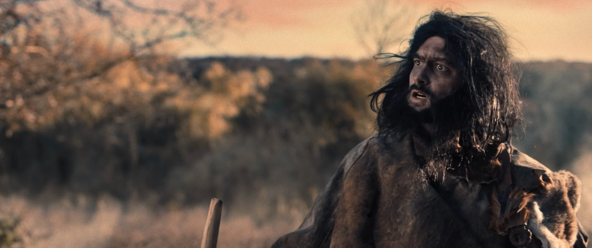 A rugged man with long, messy hair and a beard, dressed in primitive clothing, holding a small animal in his arms, standing outdoors with a blurred natural landscape in the background.