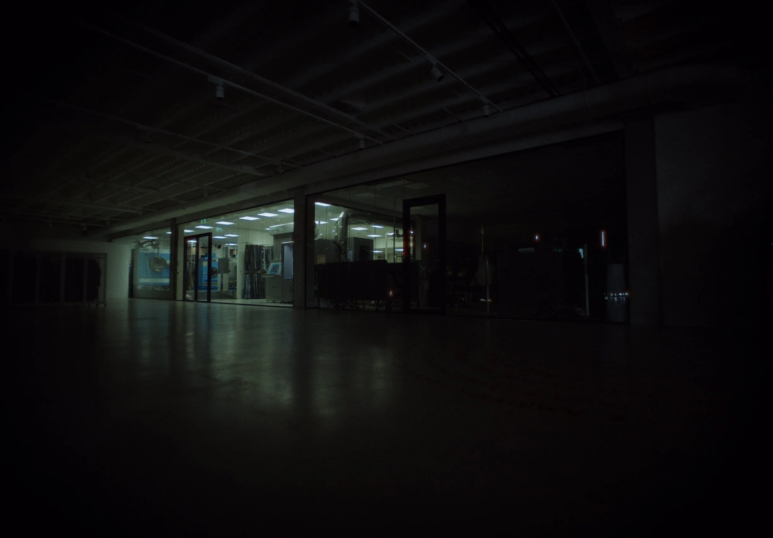 A dark interior space with a glass window showing a brightly lit room inside, possibly a gym or fitness center, with exercise equipment visible.