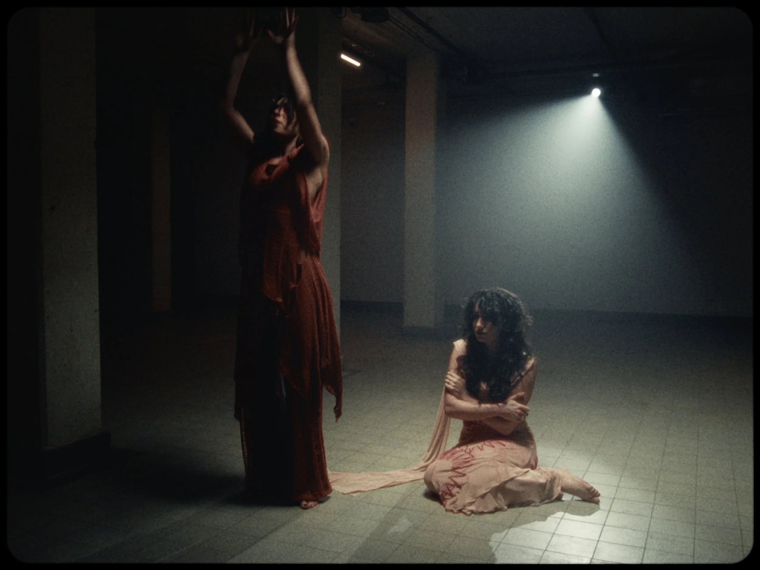 Two women in an indoor space with dim lighting and a spotlight. One woman is standing with her arms raised and hands clasped together, wearing a reddish-orange dress. The other woman is kneeling on the floor, wearing a light pink dress with red patte