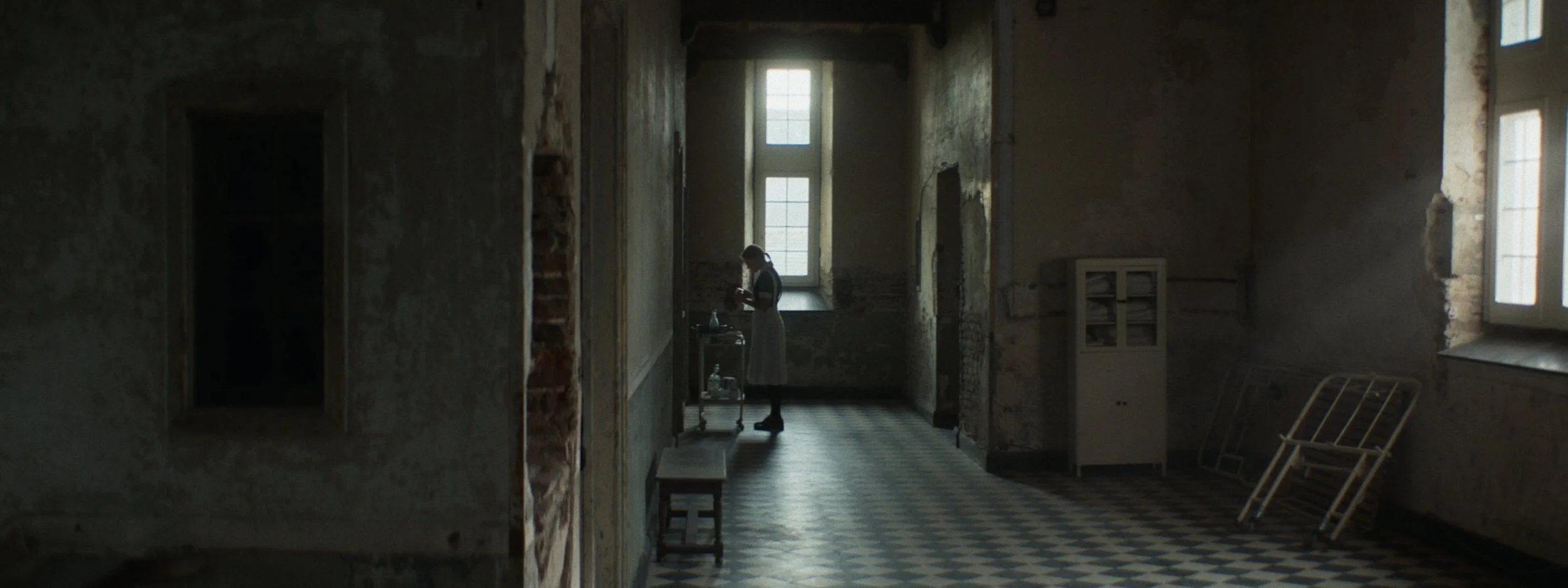 A woman in a white dress standing in a dim, old, and abandoned building with peeling walls, large windows, a checkered floor, and some furniture including a cart and a crib.