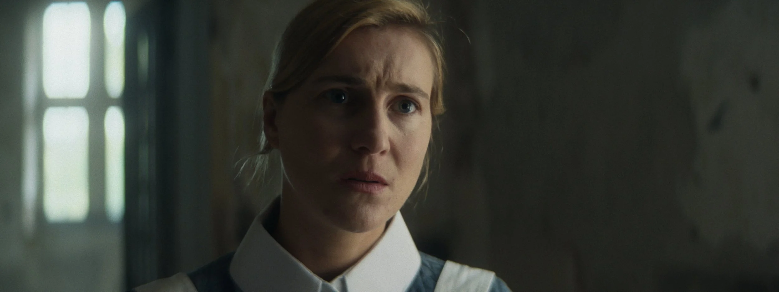 Close-up of a woman with blonde hair and blue eyes looking distressed, indoors with muted lighting, blurred background.