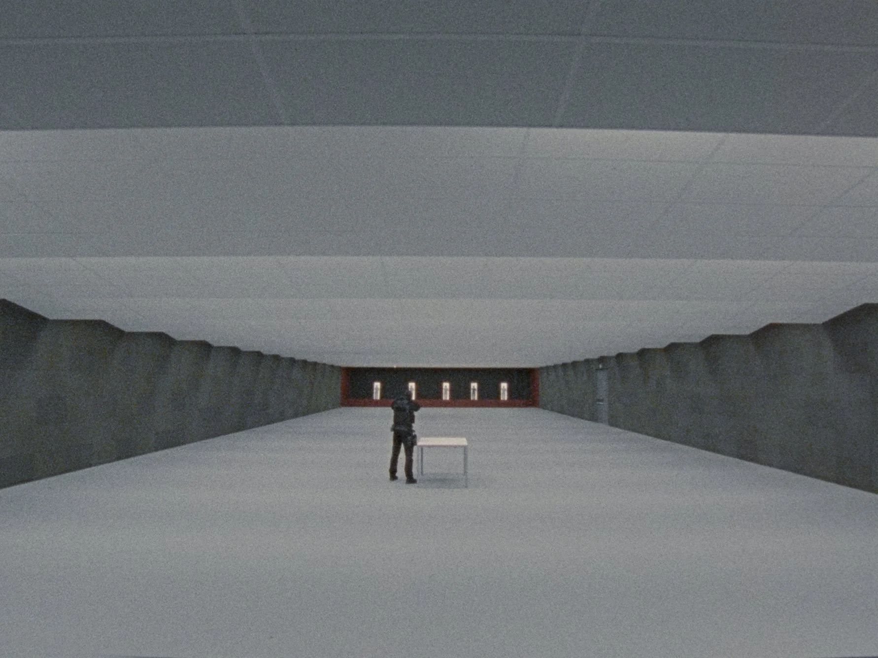 A person standing in the middle of a large, modern, and minimalist indoor space with a white floor, gray walls, and a ceiling. The person is facing away from the camera and is looking at a small table with a projection or screen on it. The area appea
