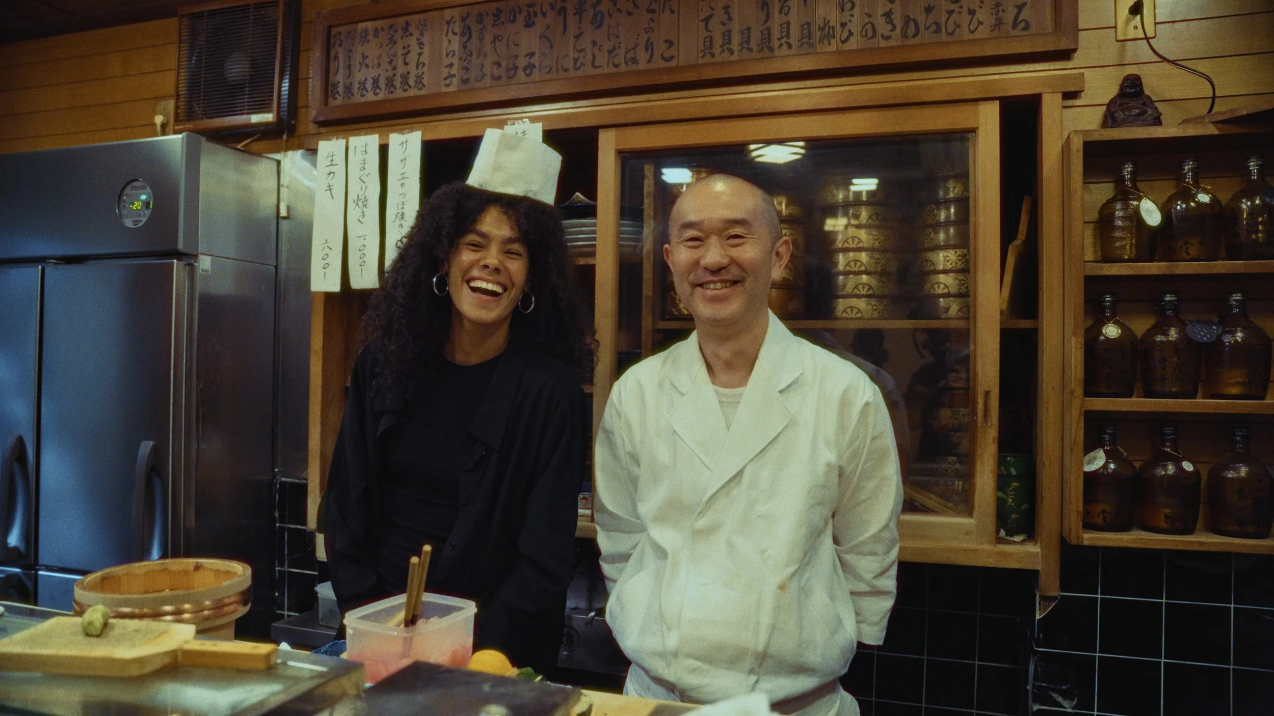 Two people, a woman with curly hair and a man in a chef's coat, smiling inside a Japanese restaurant with wooden decor and Japanese writing on the wall behind them.