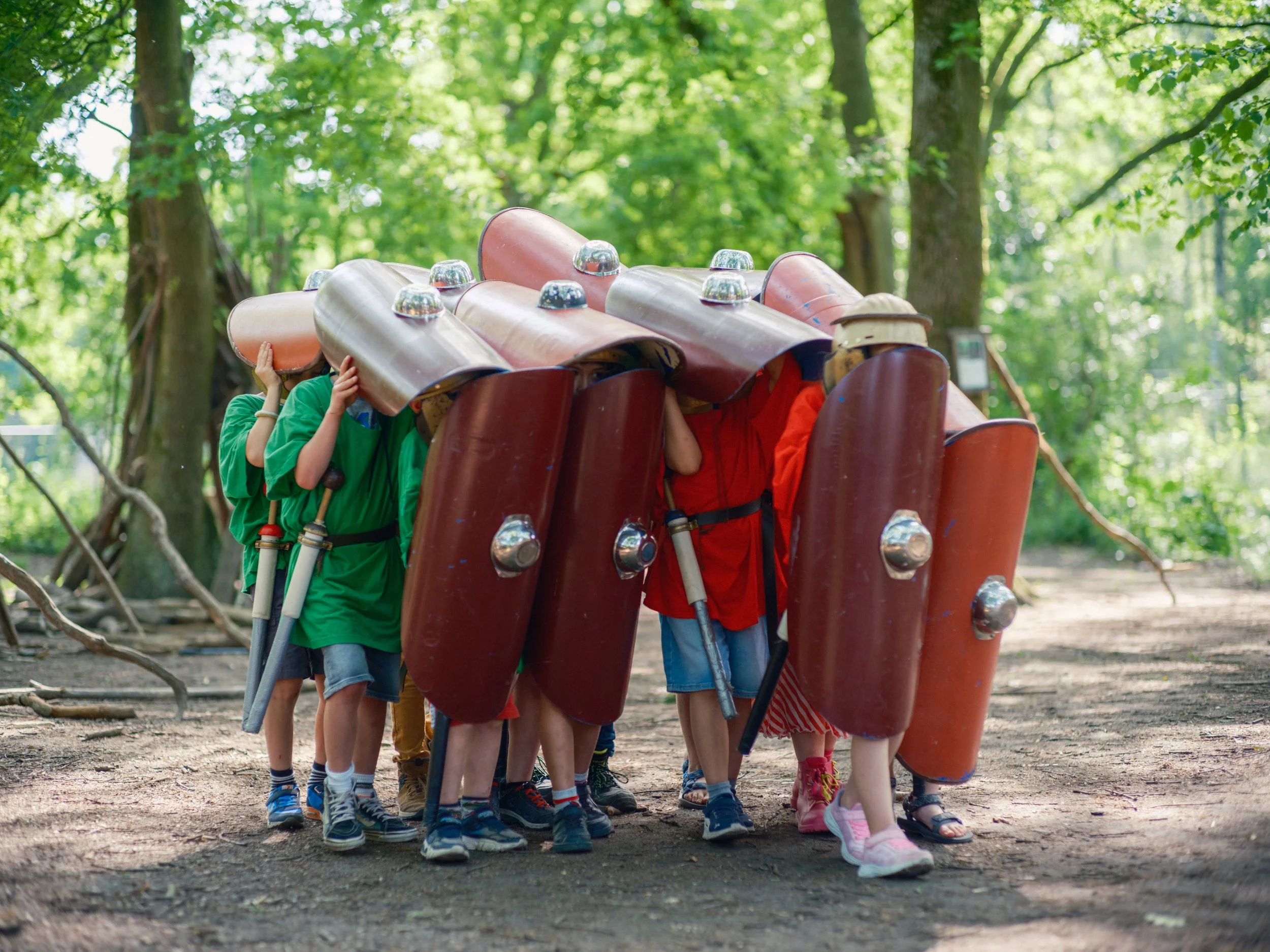 Children standing in a line, holding large foam shields while wearing helmets and colorful clothing outdoors in a park with green trees.