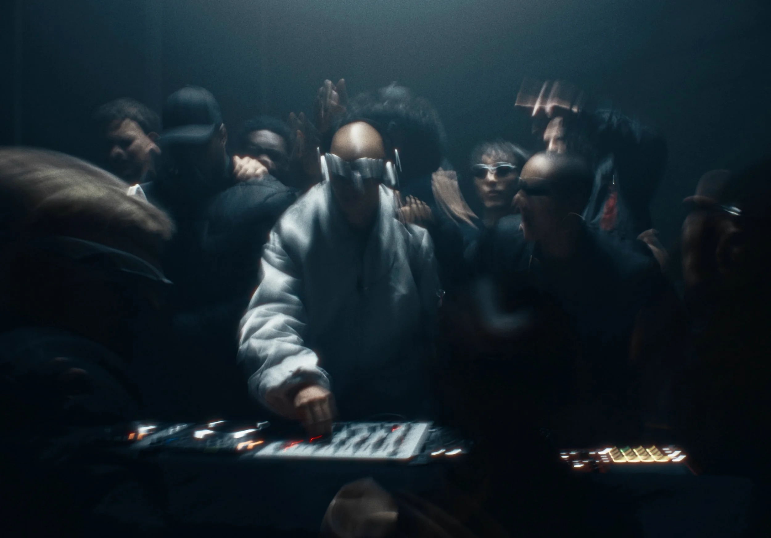 A group of people gathered around a DJ controller or sound equipment in a dimly lit room, some wearing headphones and one person wearing sunglasses, possibly at a party or music event.