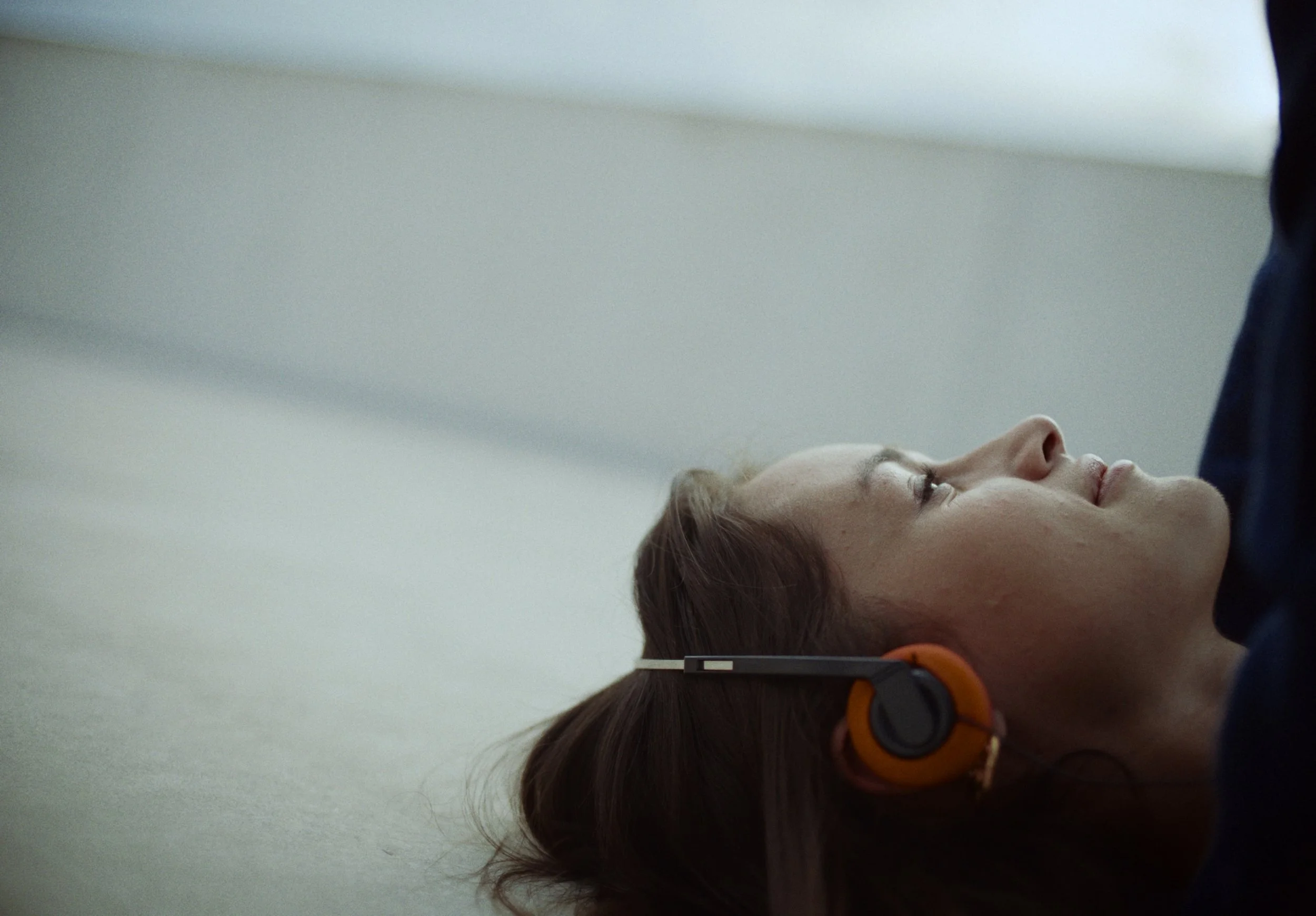 A woman is lying on the floor with her eyes closed, wearing orange headphones, and a person is standing next to her, possibly listening or talking.