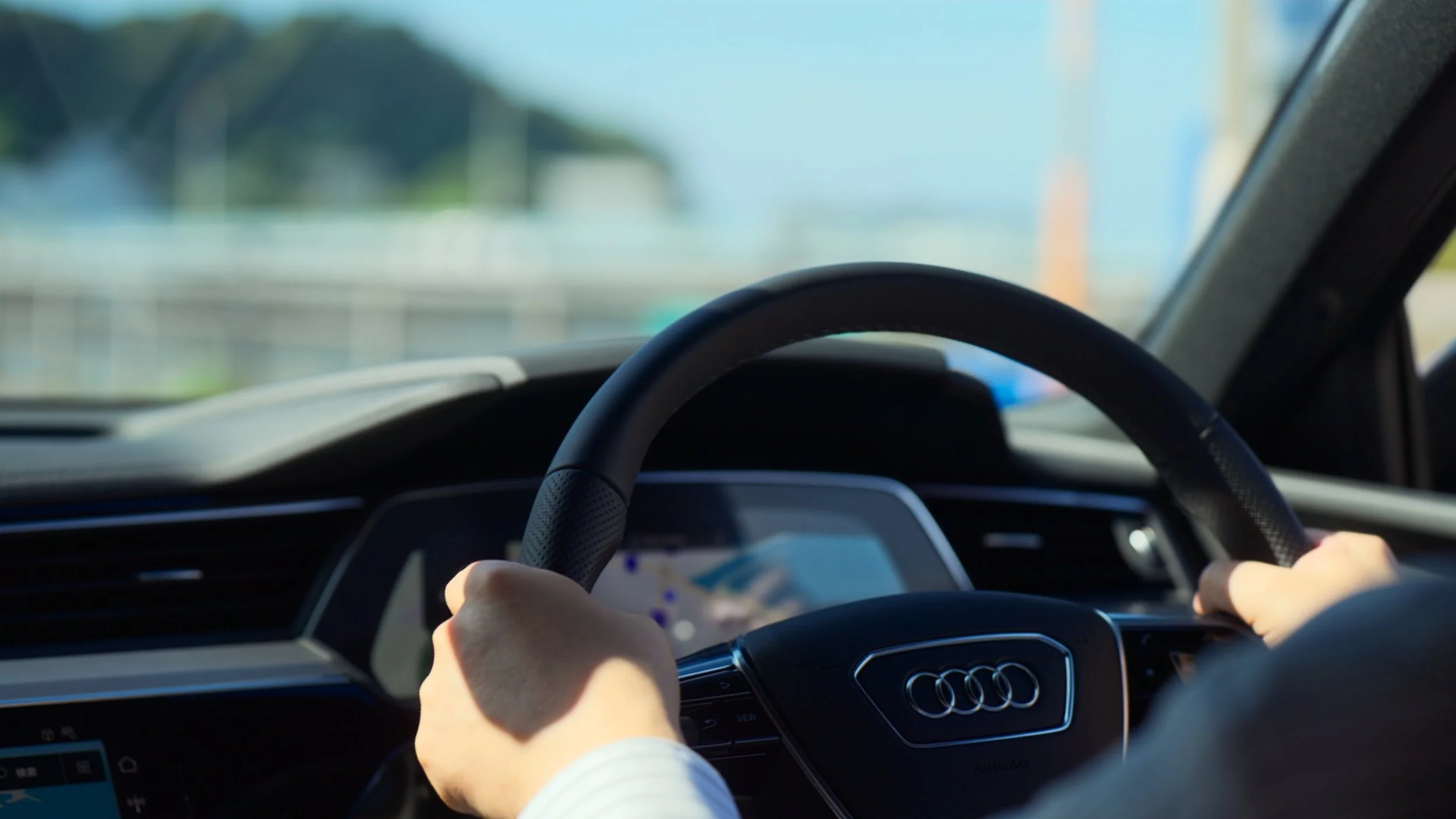 View from inside a car with a person holding the steering wheel, driving on a road with a blurred landscape through the windshield.