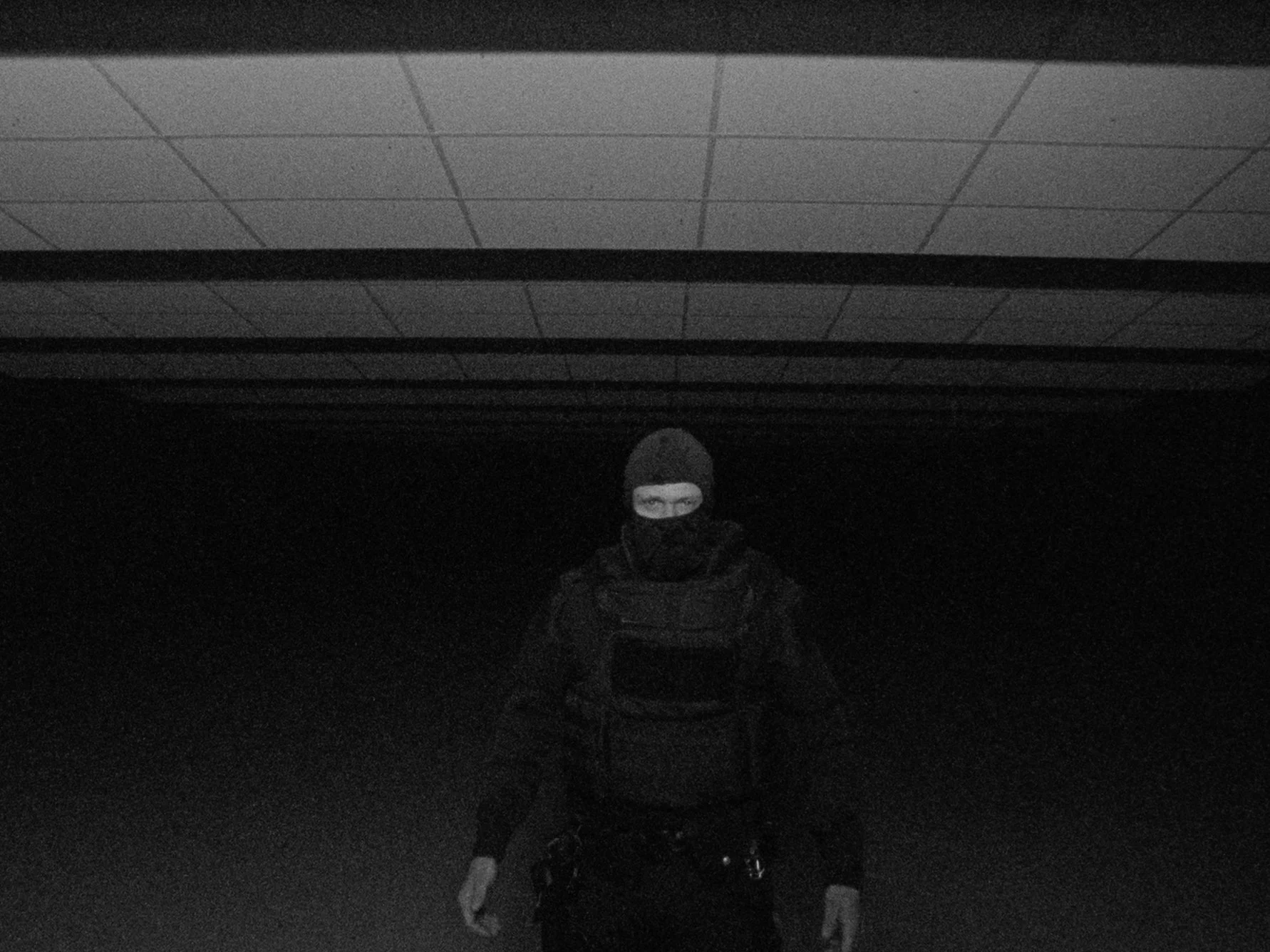 A person wearing a ski mask and dark clothing standing in a dark room or alley, with a ceiling of square acoustic tiles visible above.