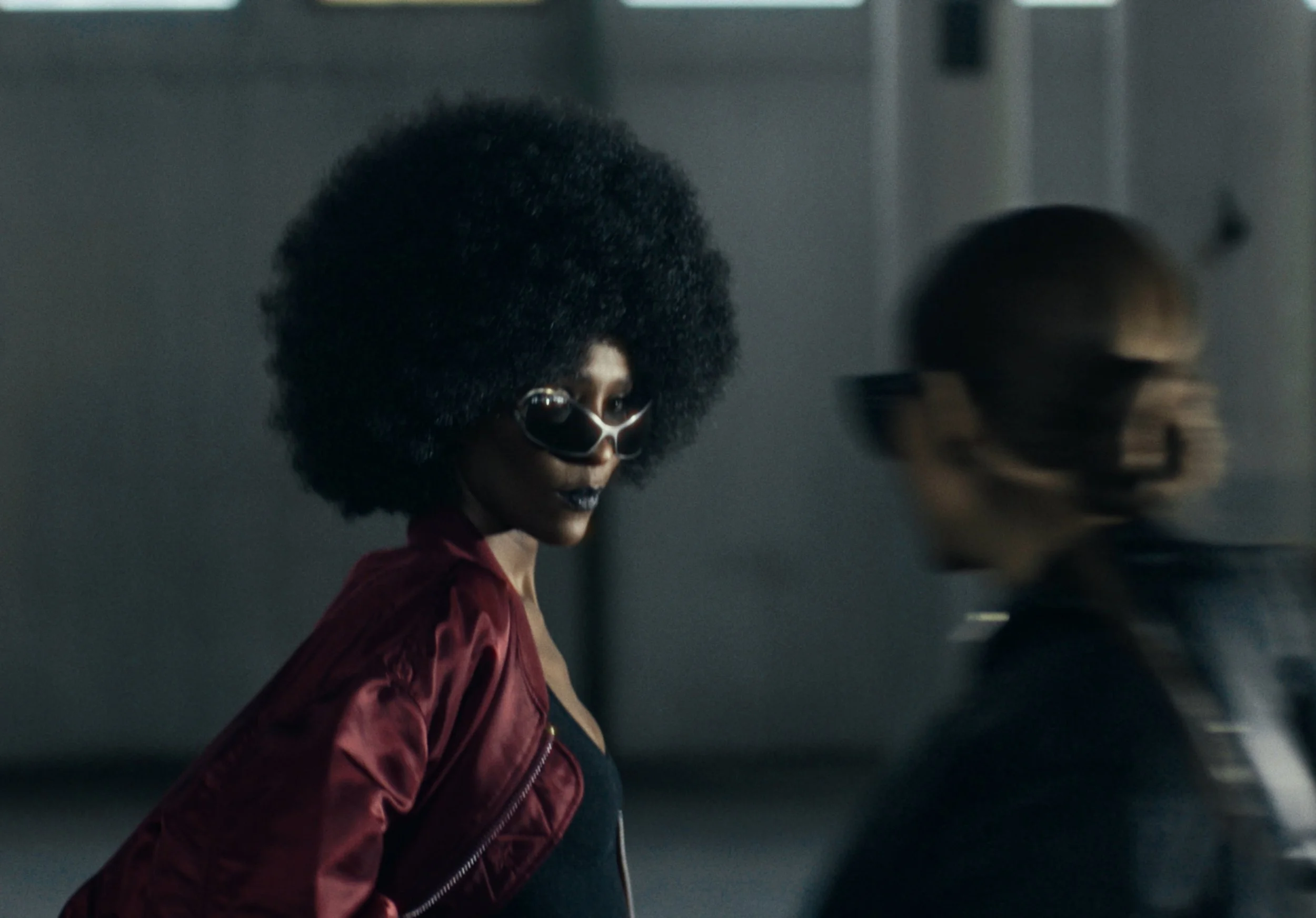A woman with dark skin, large afro hairstyle, wearing dark sunglasses and a red jacket, is walking down a corridor with a concrete wall and objects in the background.