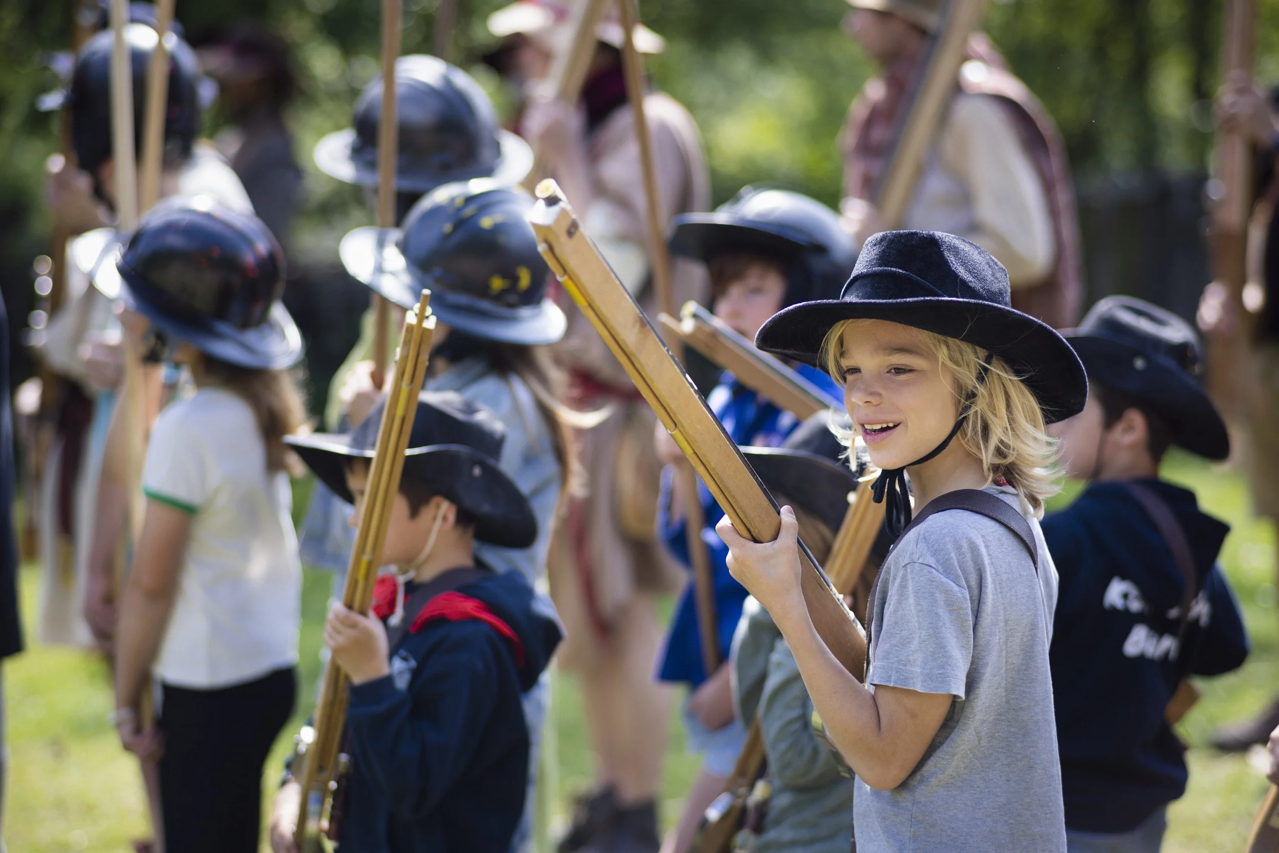 Children dressed as Pilgrims holding paddles, lined up outdoors during a reenactment or historical event, with some wearing buckled hats and traditional clothing.