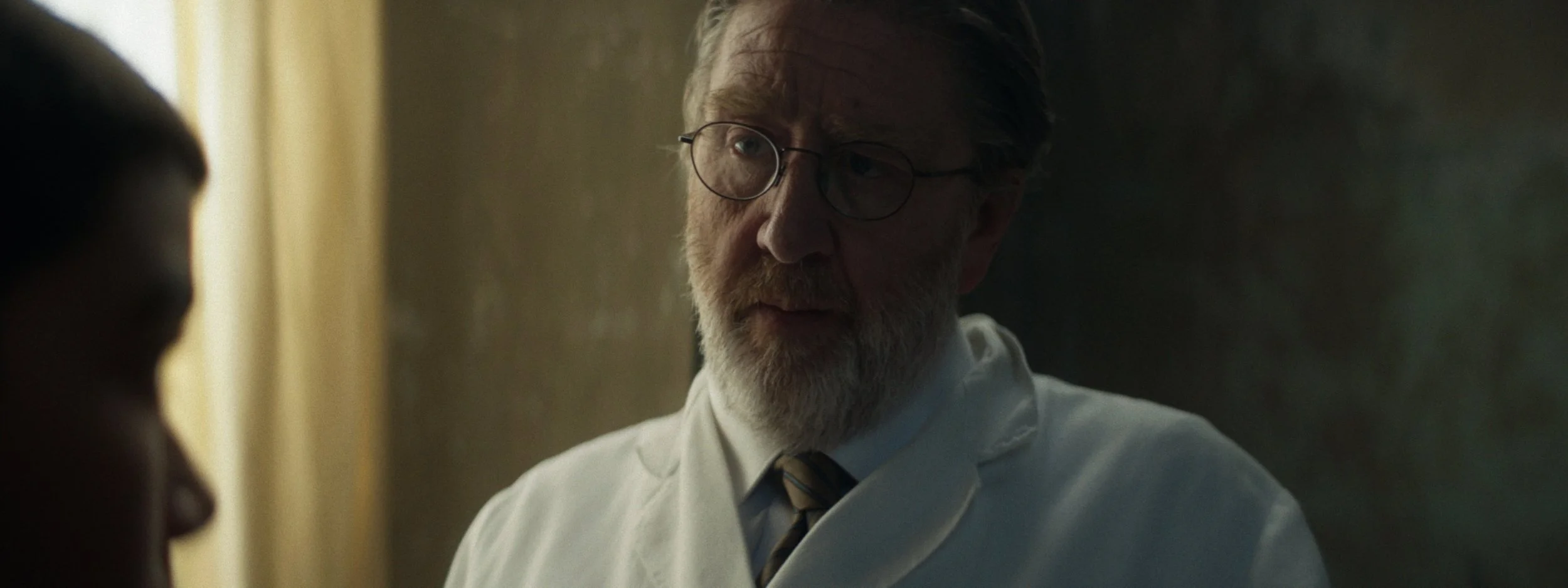 A scene from a movie depicting a middle-aged man with glasses, a beard, and gray hair wearing a white shirt and a striped tie, speaking to a young woman with dark hair.