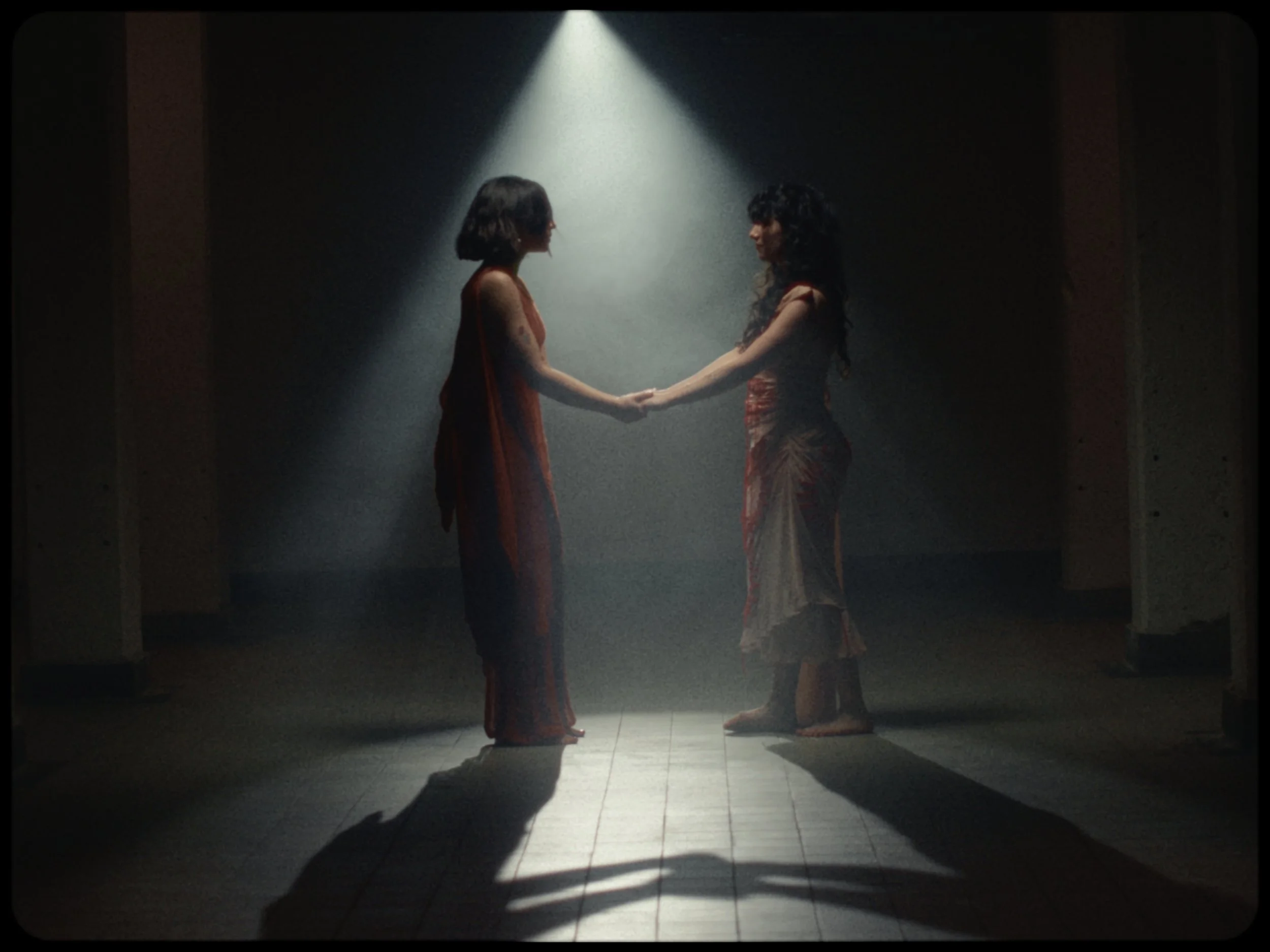 Two women holding hands and facing each other on a dimly lit stage with a spotlight overhead.