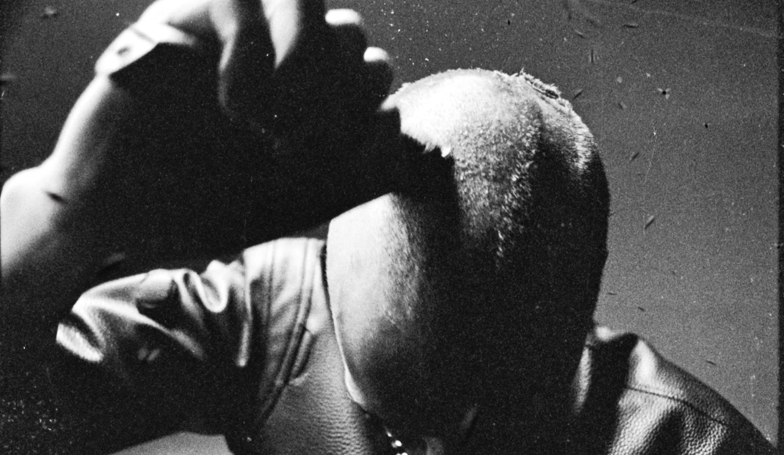 Black and white photograph of a person holding a fluffy bird, possibly a duck or chicken, close to their face. The person is wearing a jacket and gloves and is partially visible. The background is plain.