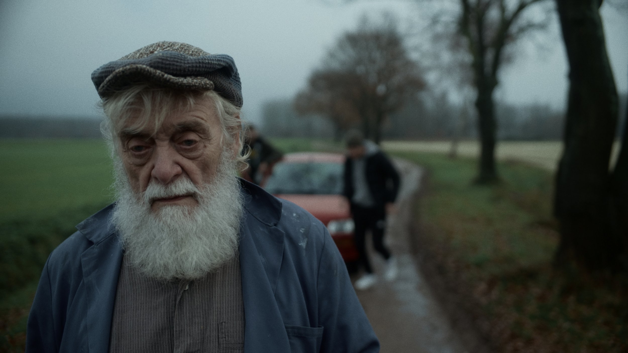 An elderly man with a white beard and gray hair wearing a layered cap and jacket stands outdoors on a cloudy, foggy day. In the background, a young man leaning against a car is seen, with trees and a grassy area along a rural road.