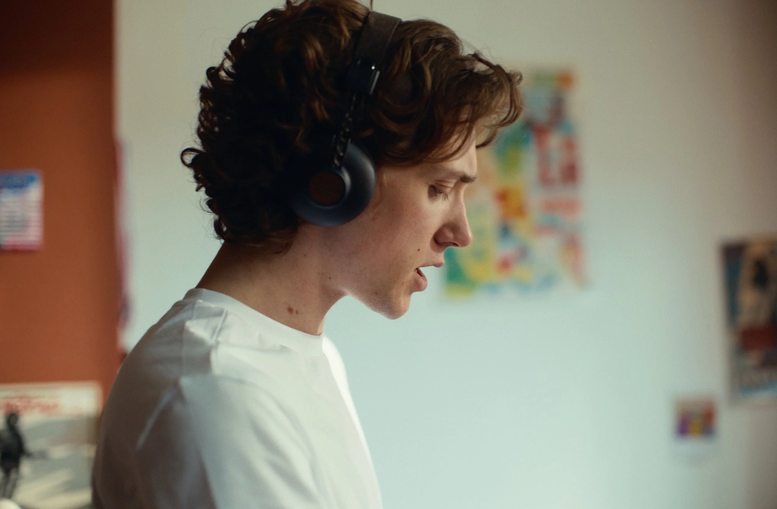 A young woman with curly brown hair wearing headphones, standing in profile in front of a white wall with colorful posters.