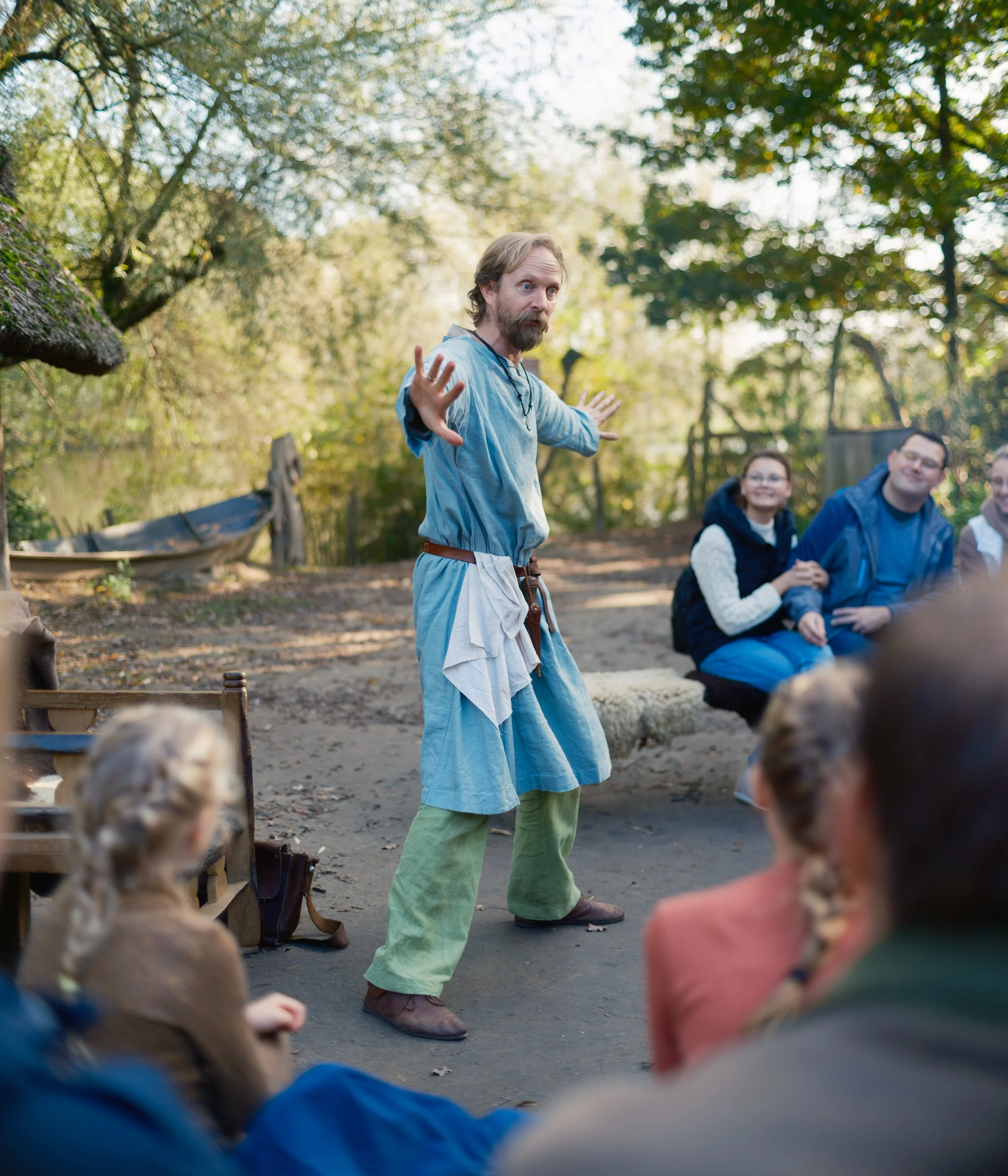 A man dressed in peasant or medieval costume, with a blue tunic and green pants, is pointing and speaking to a group of children outdoors in a wooded area with sunlight filtering through the trees.