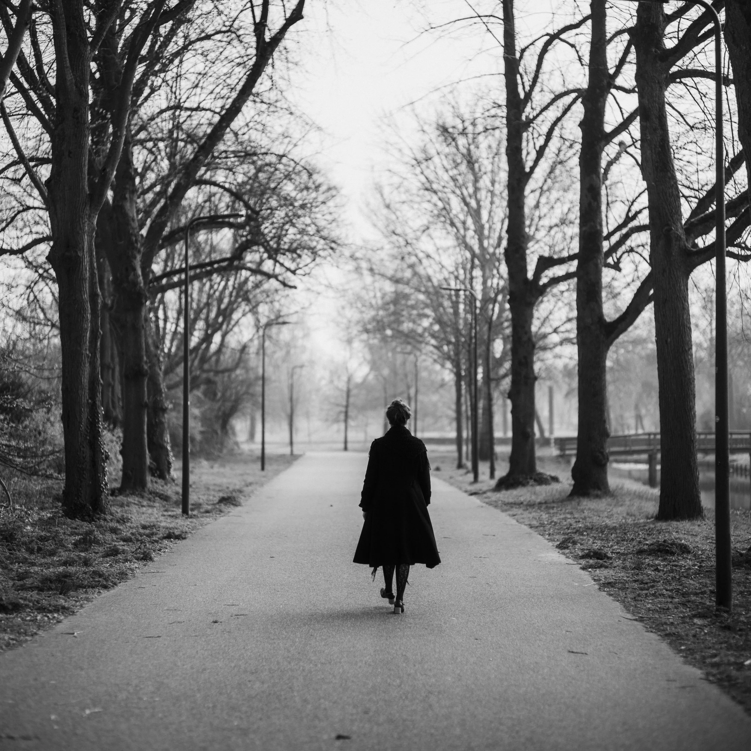 A black and white photo of a person walking on a paved path in a park lined with tall, leafless trees, with street lamps along the pathway.