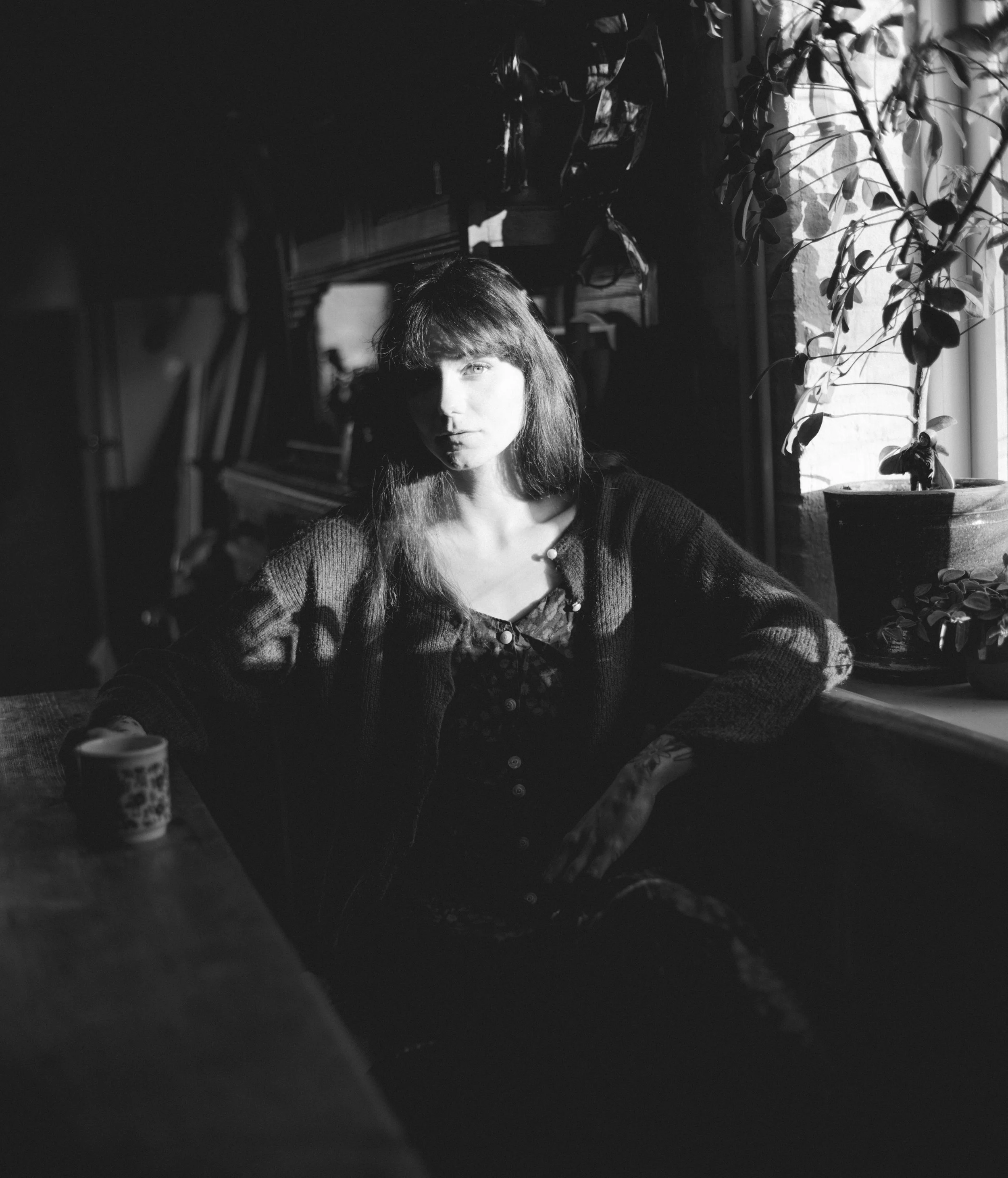 Black and white photo of a woman with long hair, sitting near a window with sunlight, plants on the windowsill, and furniture in the background.
