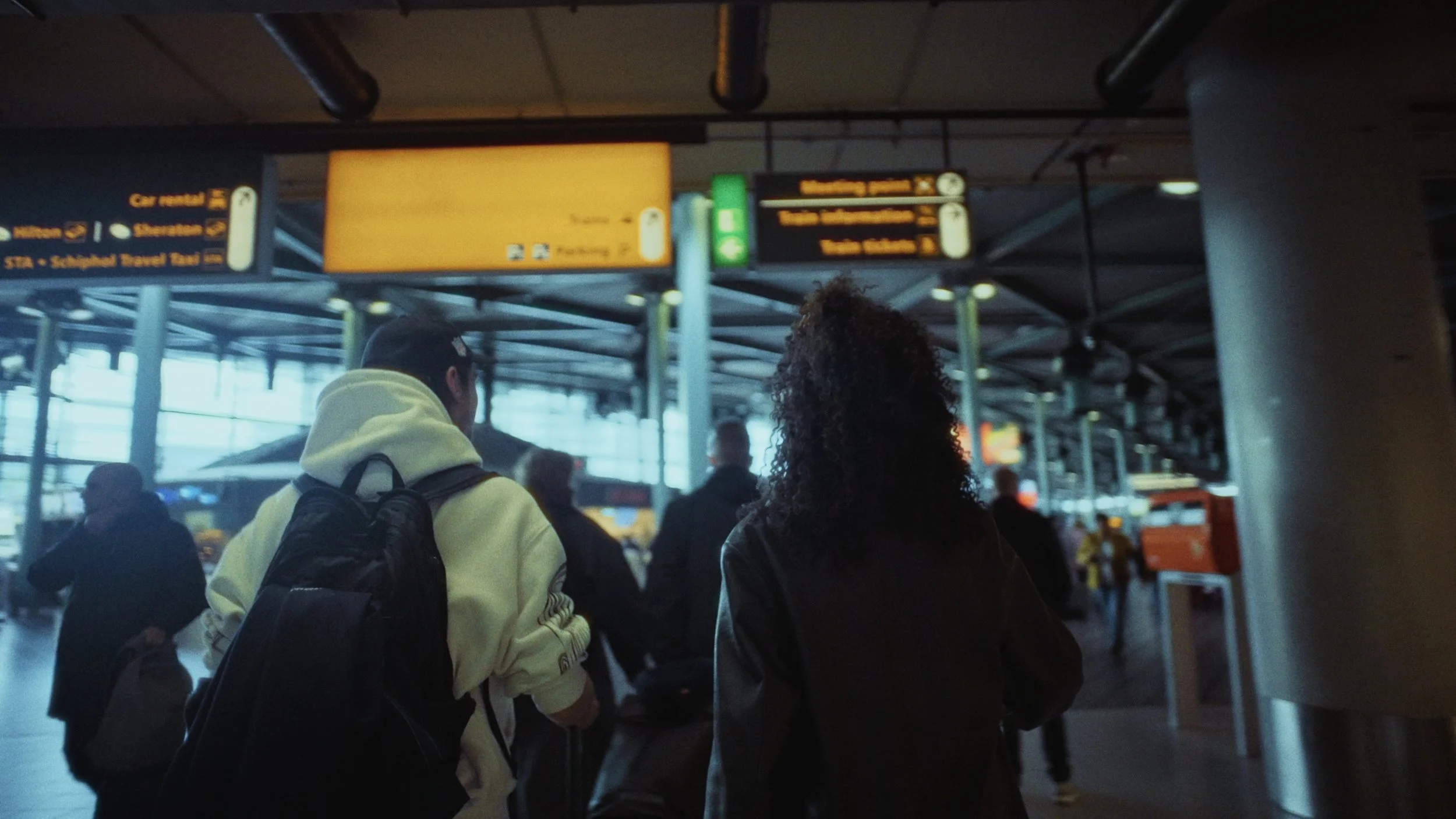 People standing in line at an airport terminal, with overhead signs for car rental, transportation, and other services, and large windows in the background.