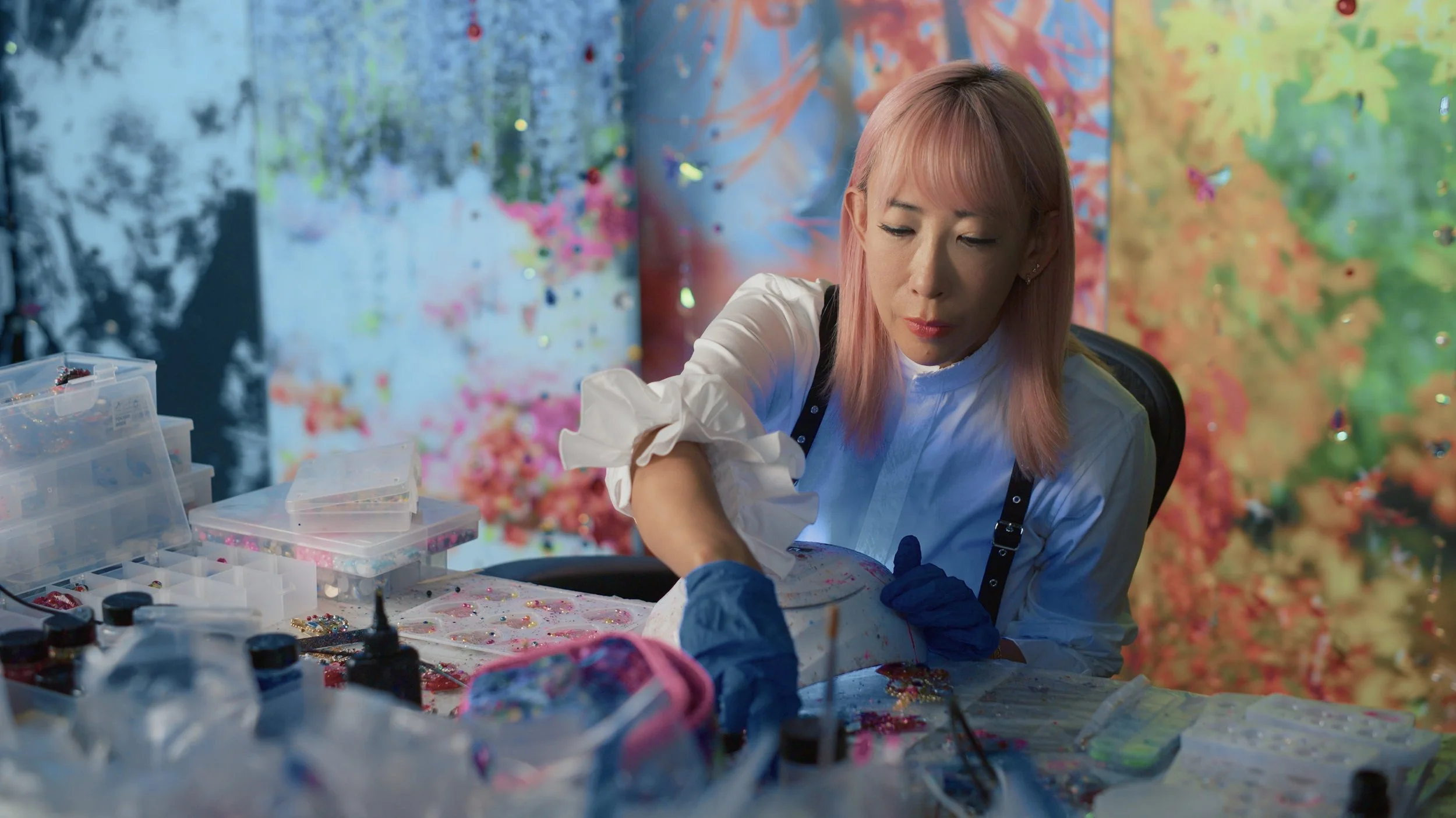 A woman with pink hair working on a craft in a colorful art studio filled with paint and glitter.