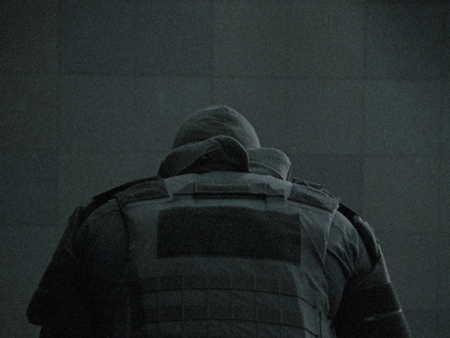 A person dressed in tactical gear, including a helmet and a backpack, seen from behind in a dark setting.