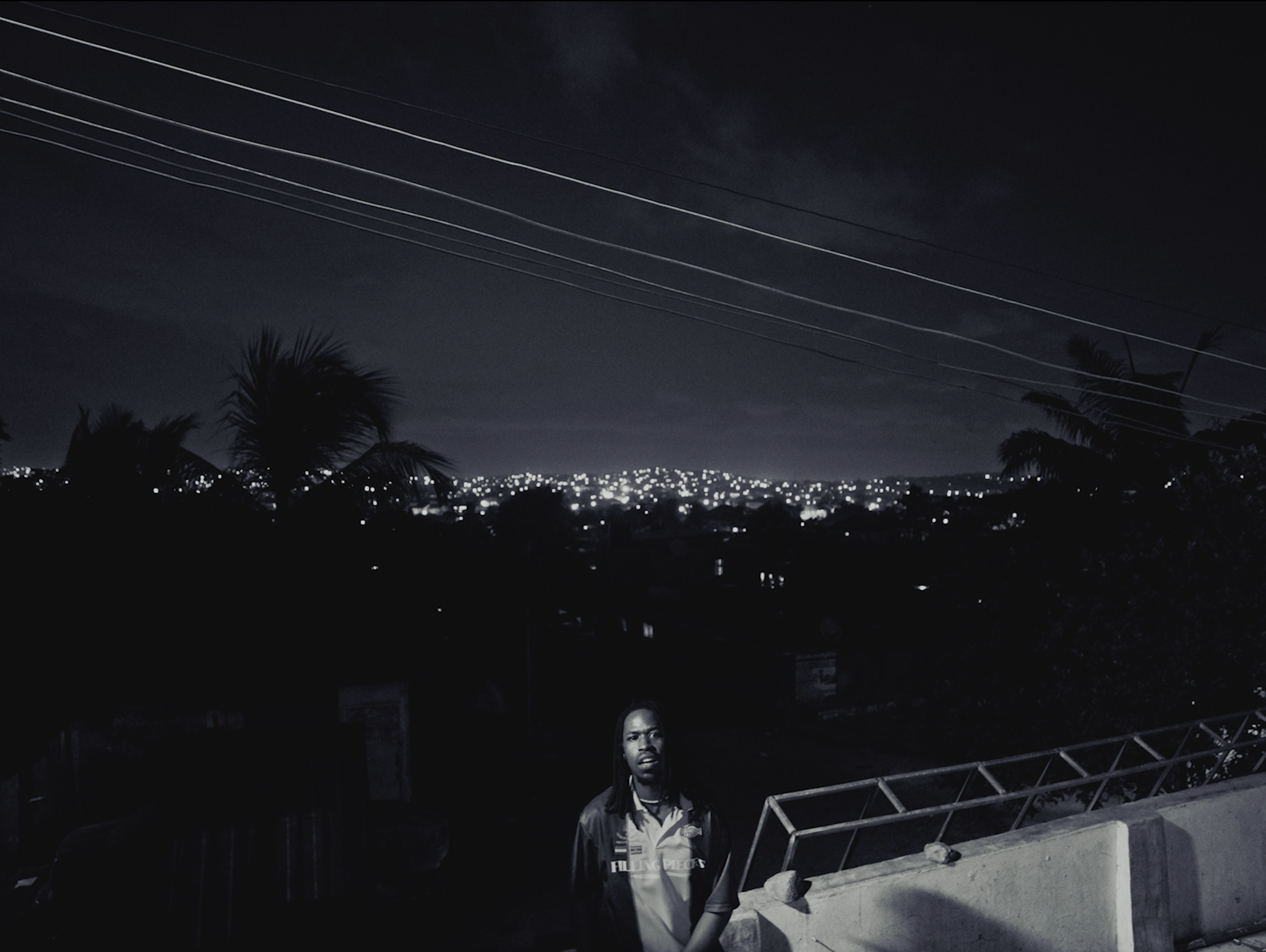 Nighttime scene with power lines crossing the dark sky, city lights in the distance, and a man standing near a railing in the foreground.
