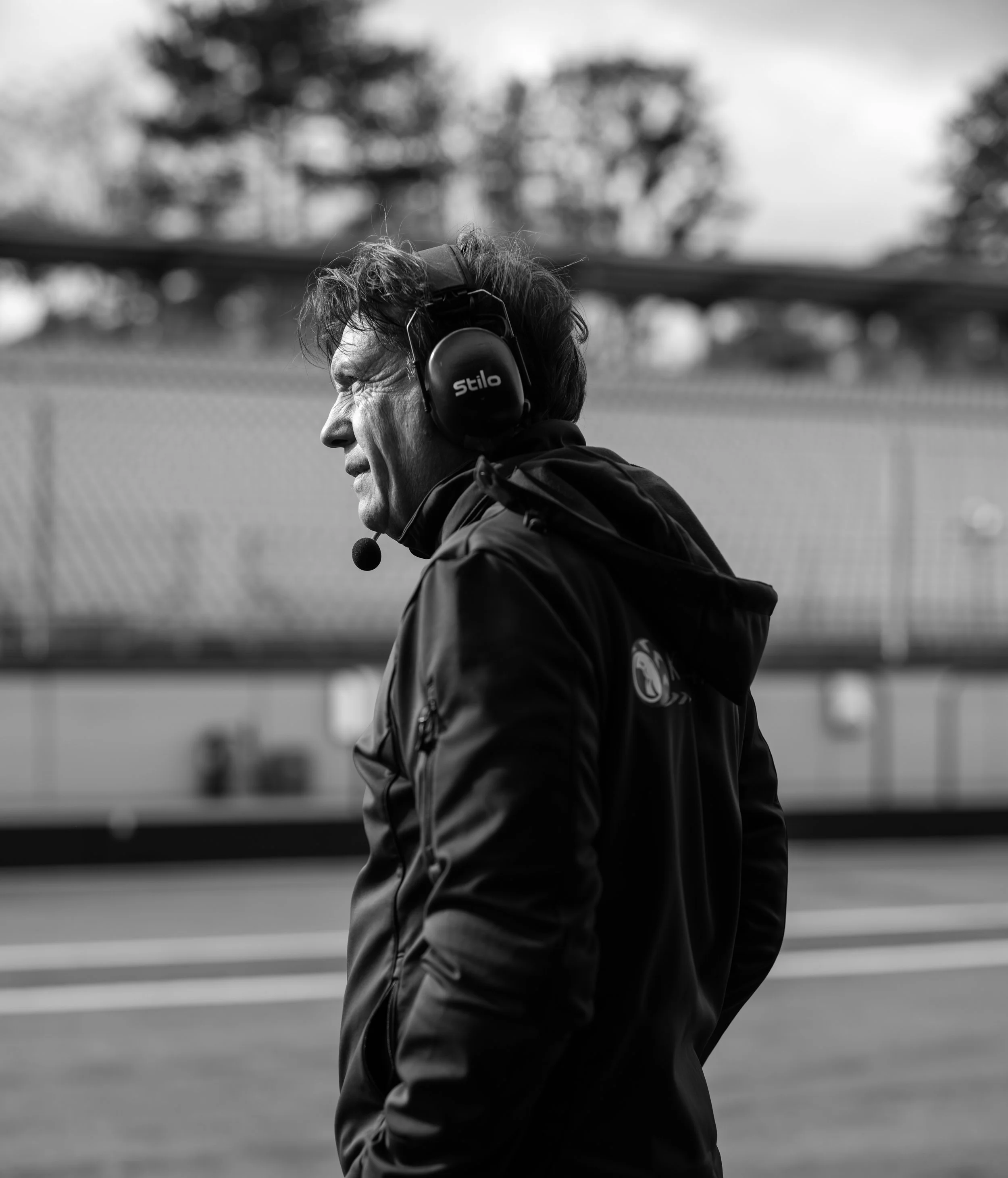 A black and white photo of a man wearing a headset and dark jacket, walking outside with blurred trees and a fence in the background.