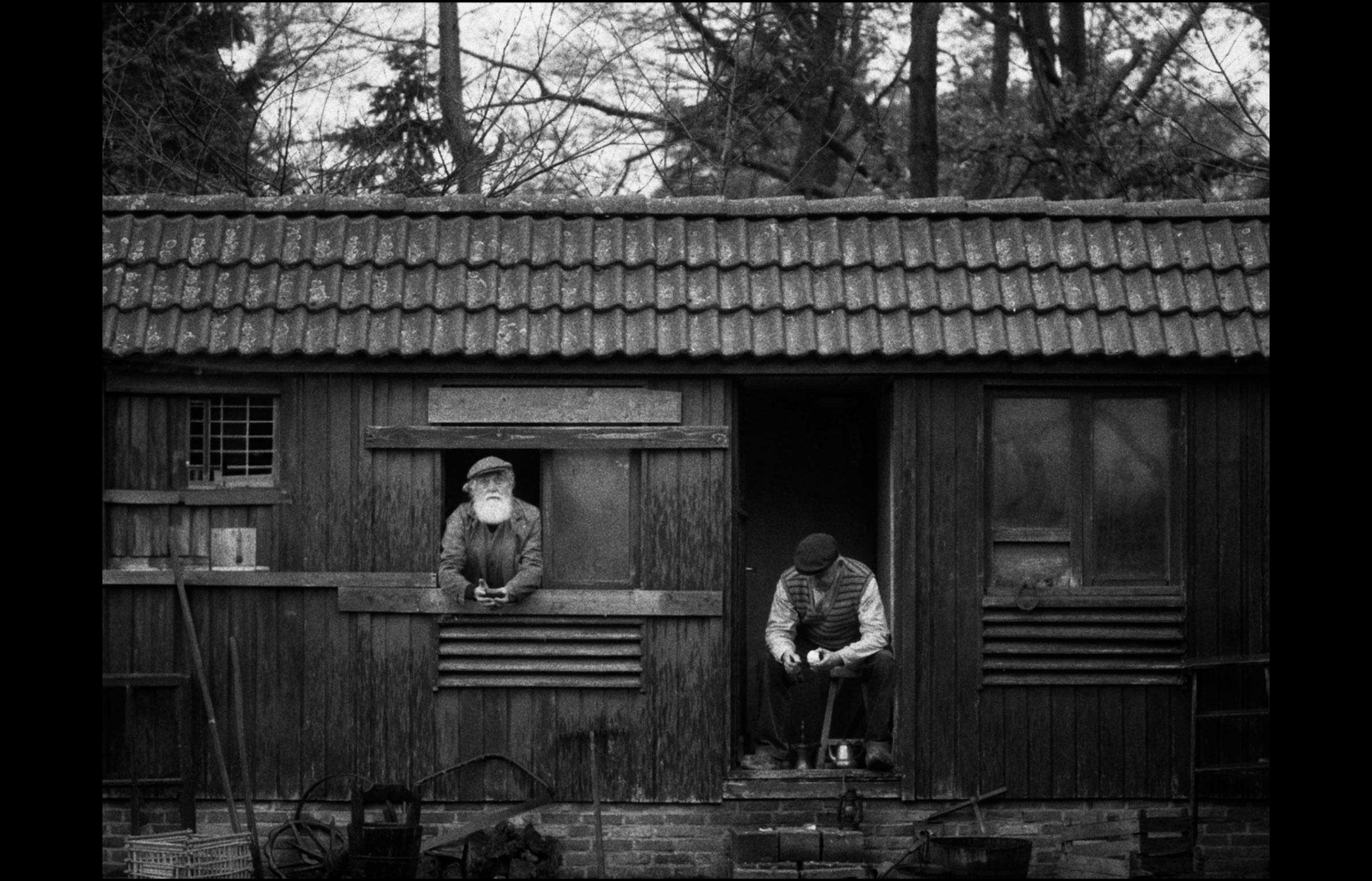 Two men are standing in front of a wooden house with a small window and a door. One man is leaning on the windowsill, holding a cigarette, and wearing a cap and jacket. The other man, sitting on a ledge below the window, is looking down and also wearing a cap and jacket. The house has a tiled roof, and there are trees in the background. The photo is in black and white.
