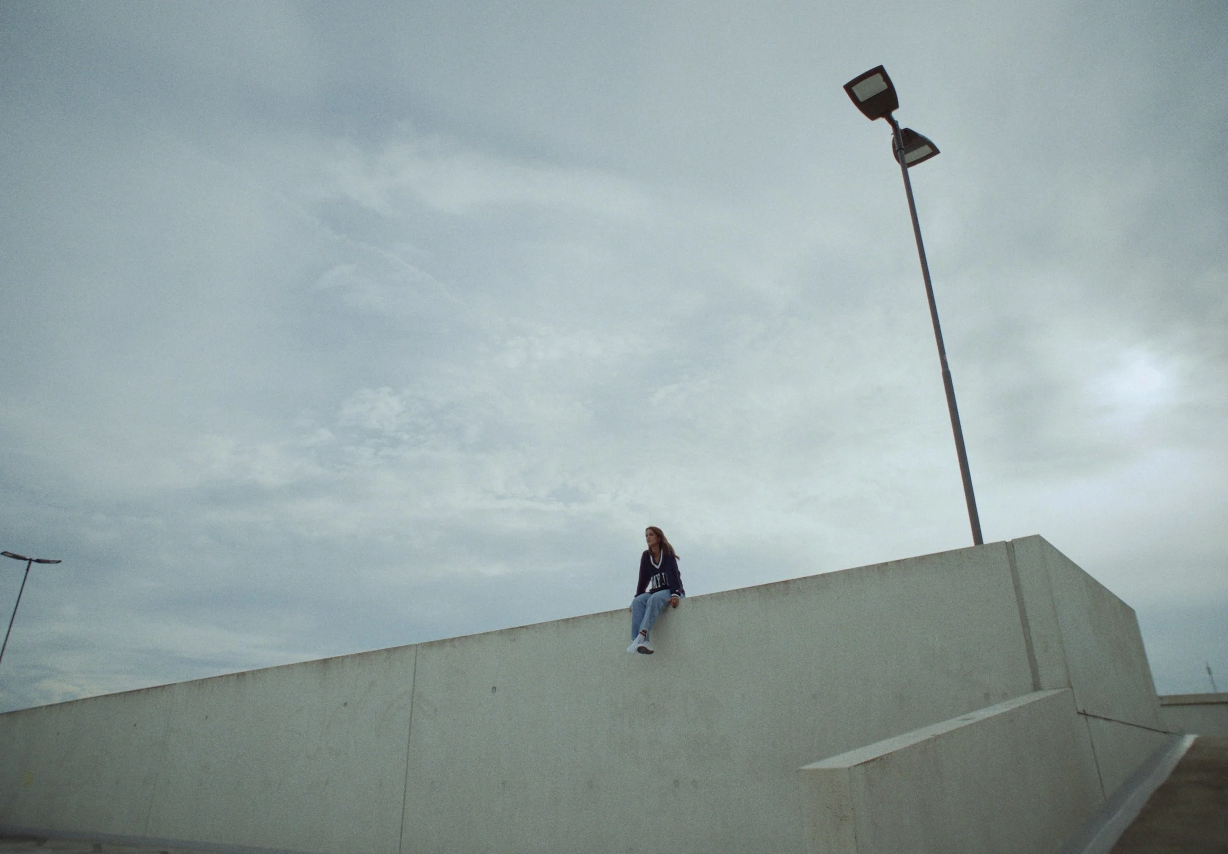 A woman with a hoodie sitting on a ledge outdoors under a cloudy sky, with streetlights nearby.