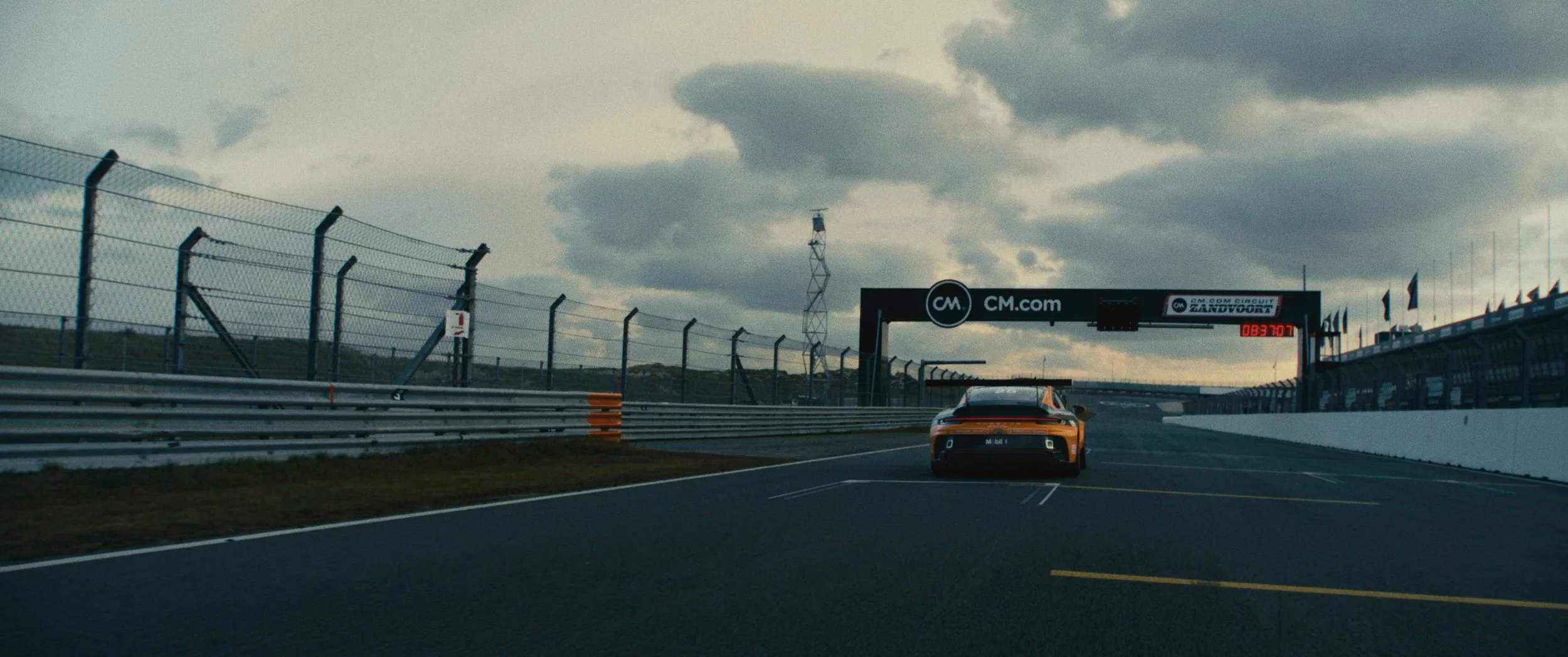 A race car is driving on a track with a digital clock and a bridge overhead. The sky is cloudy.