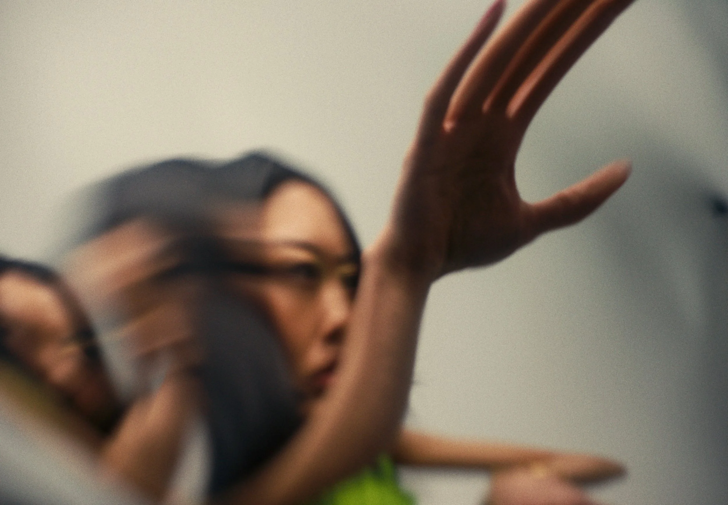 Close-up of a person's face and hand, with the person's face blurred and the hand raised and in focus, against a plain background.