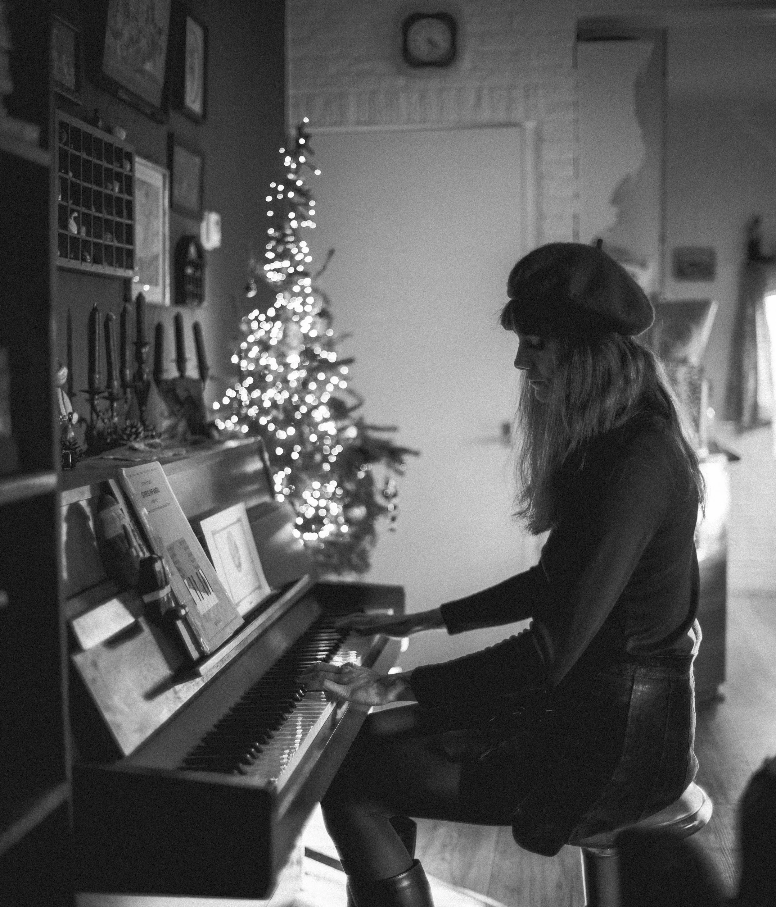 A woman sitting at a piano playing keys, with a decorated Christmas tree in the background. The scene is in black and white.