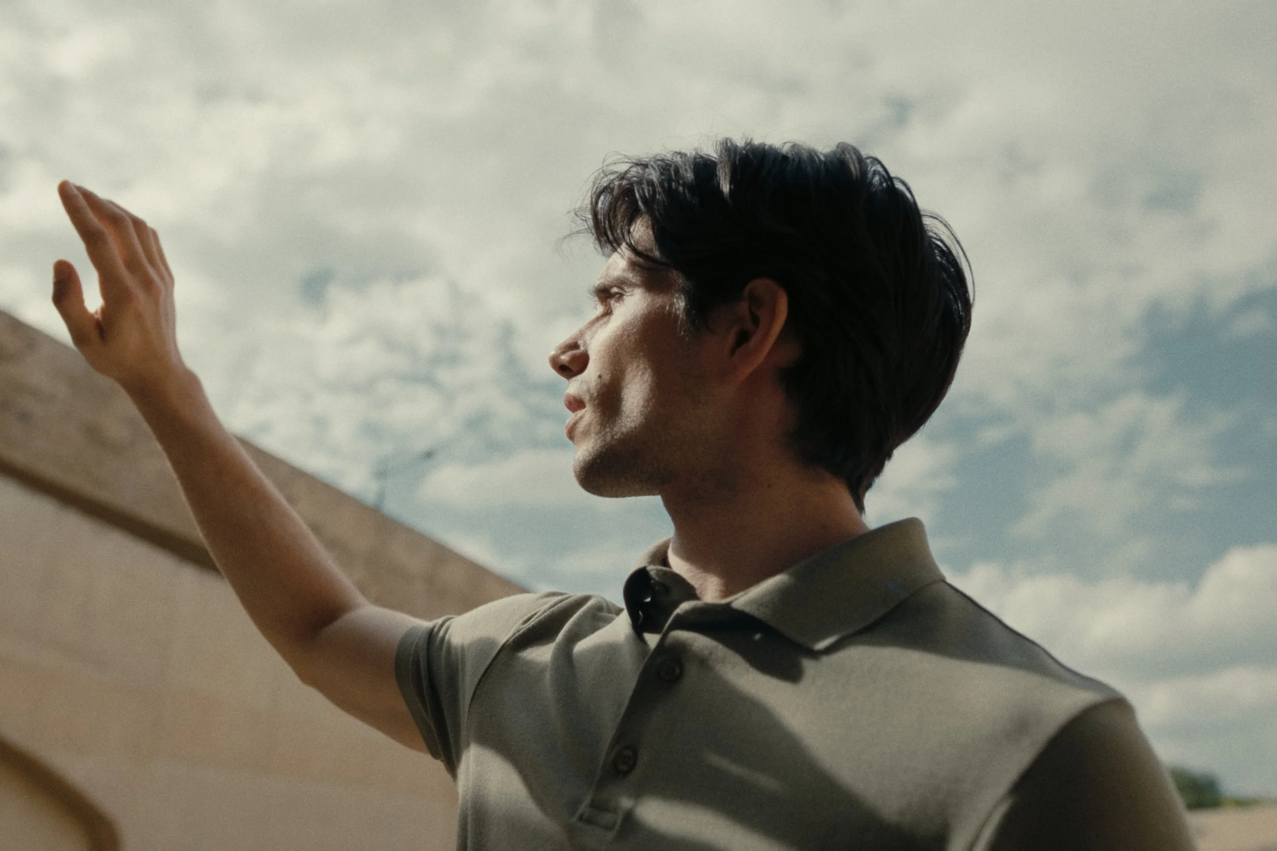 A man with dark hair wearing a gray polo shirt, standing outdoors under cloudy skies, reaching out with his hand, possibly touching a wall or surface.