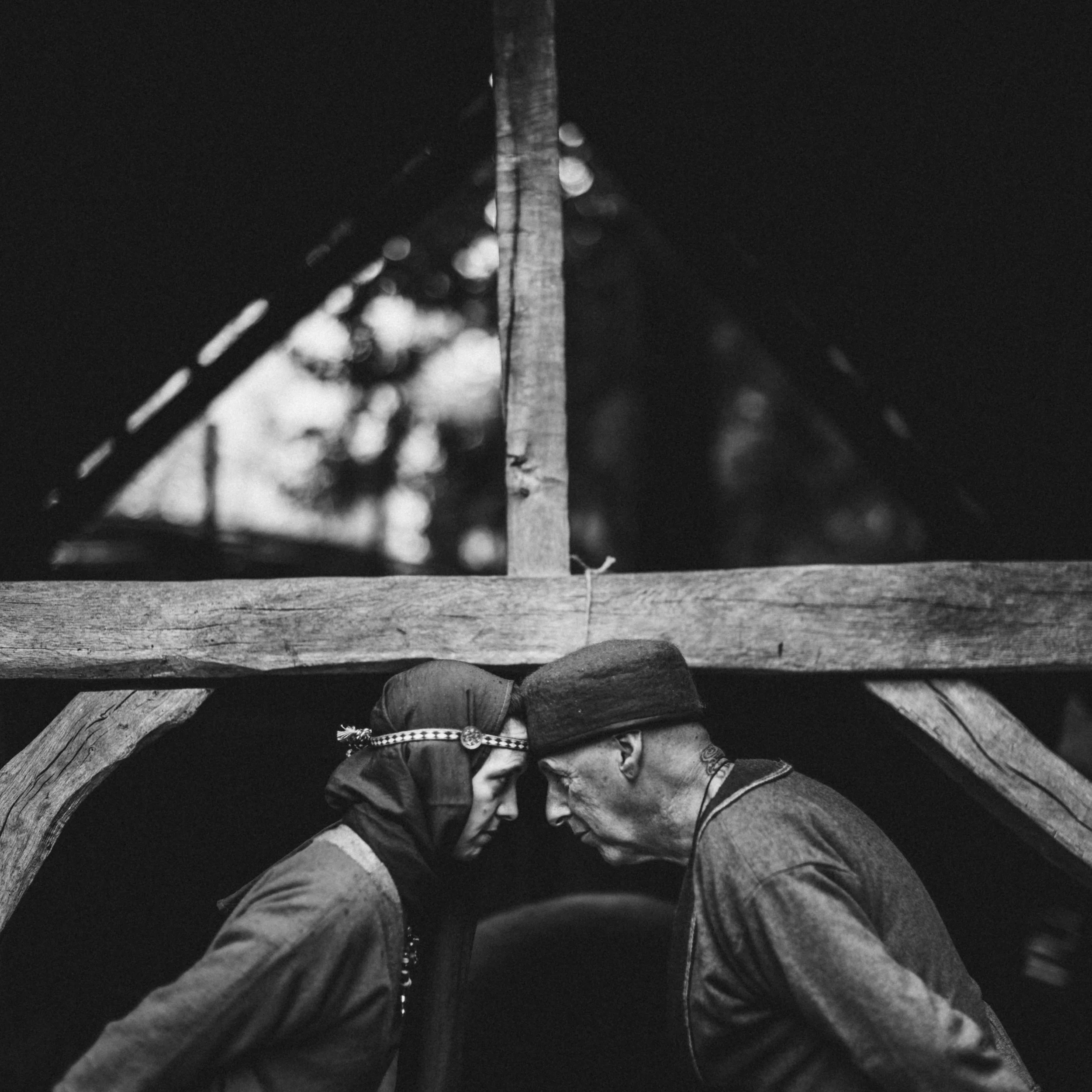 A black and white photo taken from a low angle showing a person sitting on the ground with their back against a wooden structure, wearing a jacket and a patterned lanyard or badge, with a chain link fence nearby and small raindrops or water droplets 