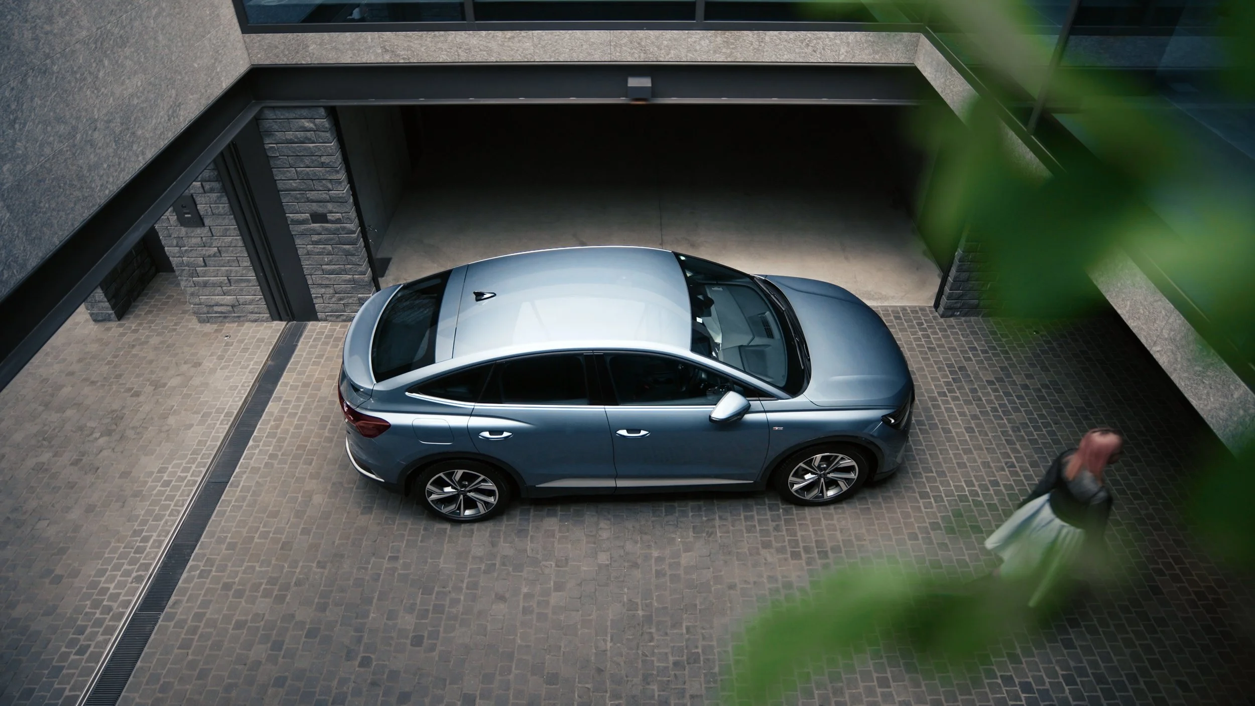 A silver SUV parked in a driveway outside a modern building with brick and concrete walls, partially obscured by a blurred green plant in the foreground.