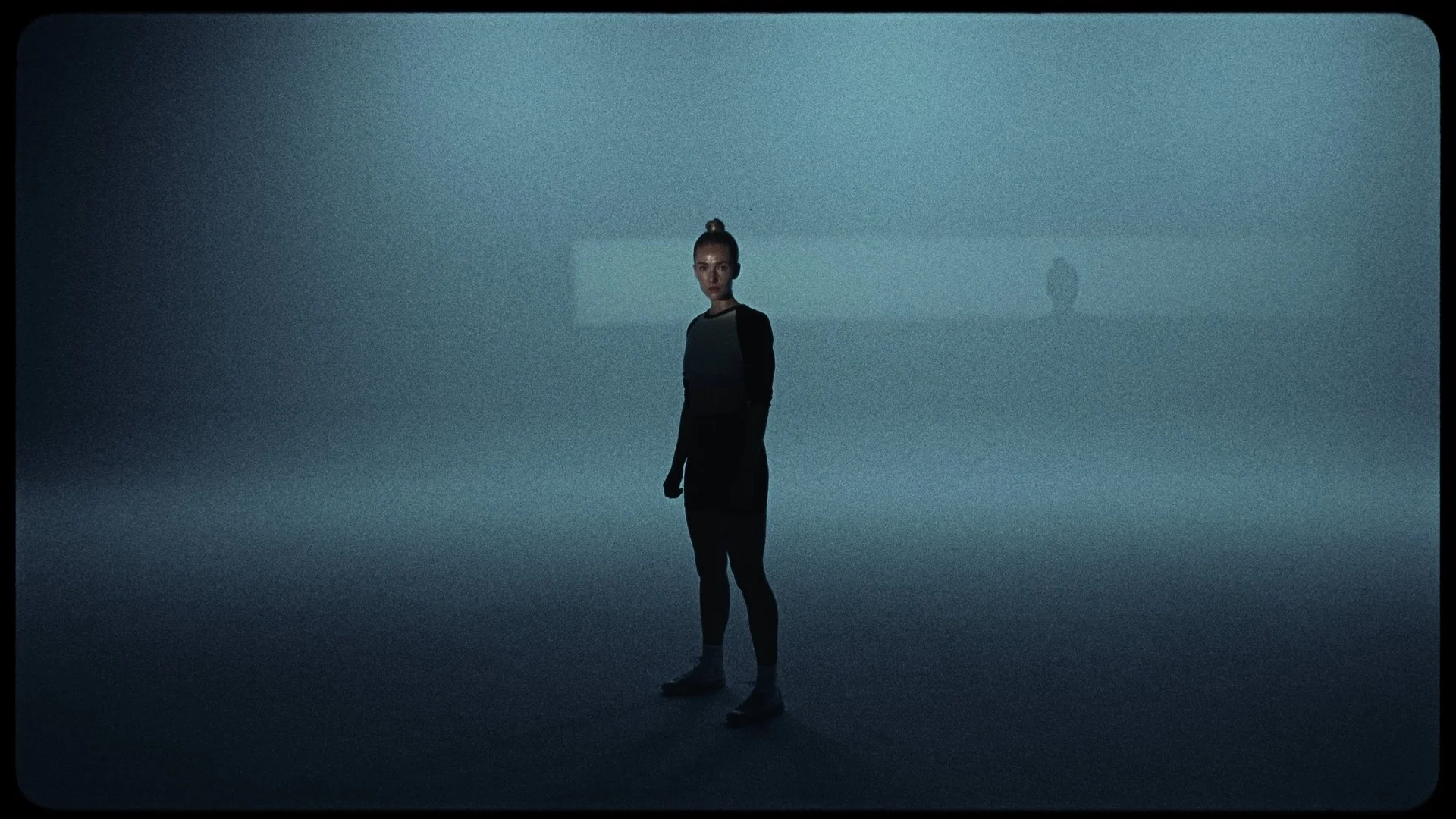 A woman with a high bun hairstyle standing in a dimly lit room with a foggy or smoky atmosphere. She is wearing a black and gray long-sleeve top and black pants. There is a faint shadow of her on the wall behind her, and the floor appears reflective.